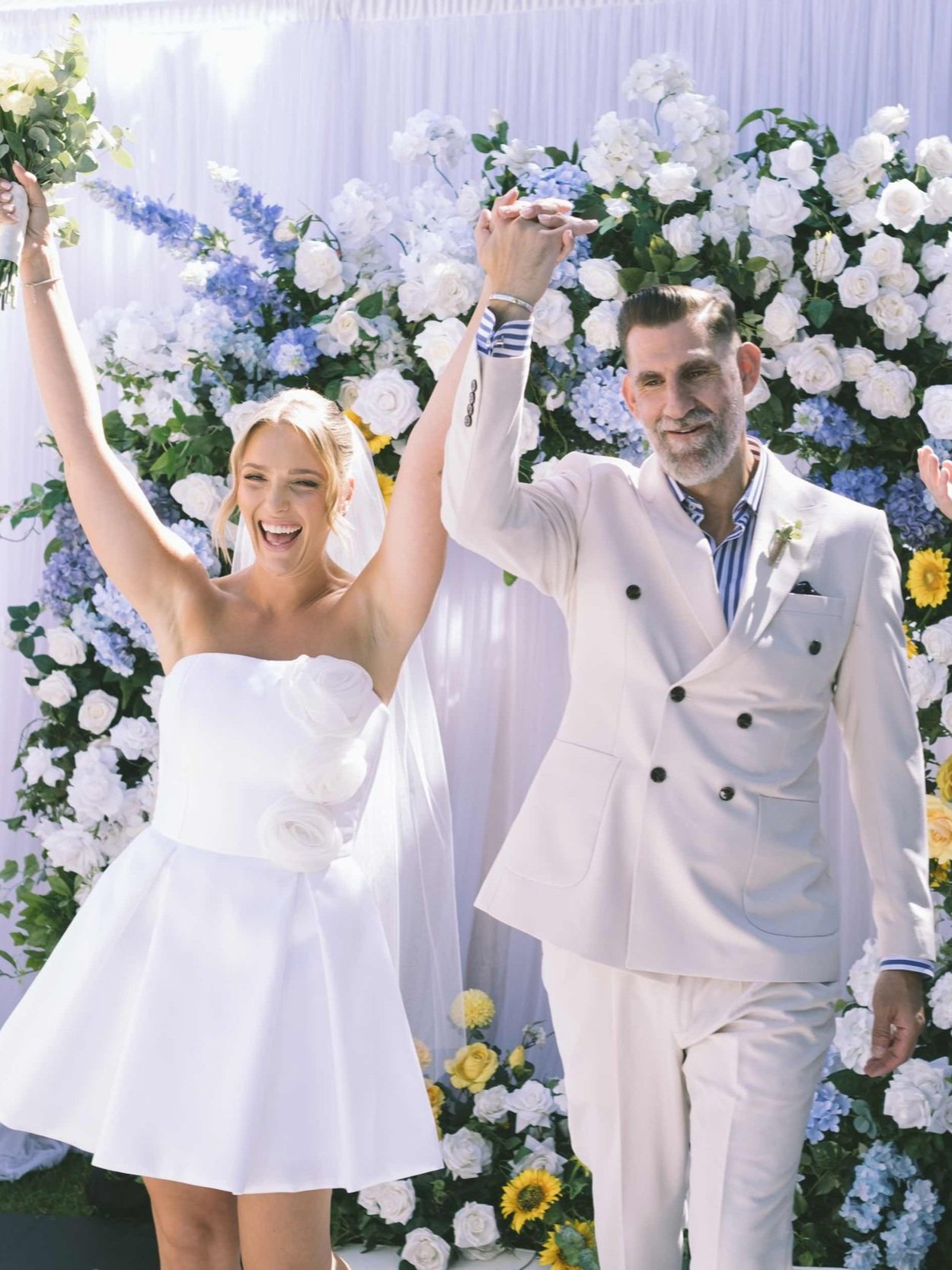A happy couple holding hands in front of a floral backdrop. The woman is wearing a short white dress with floral accents, and the man is in a light-colored suit. Both appear to be celebrating or at a special event, like a wedding.