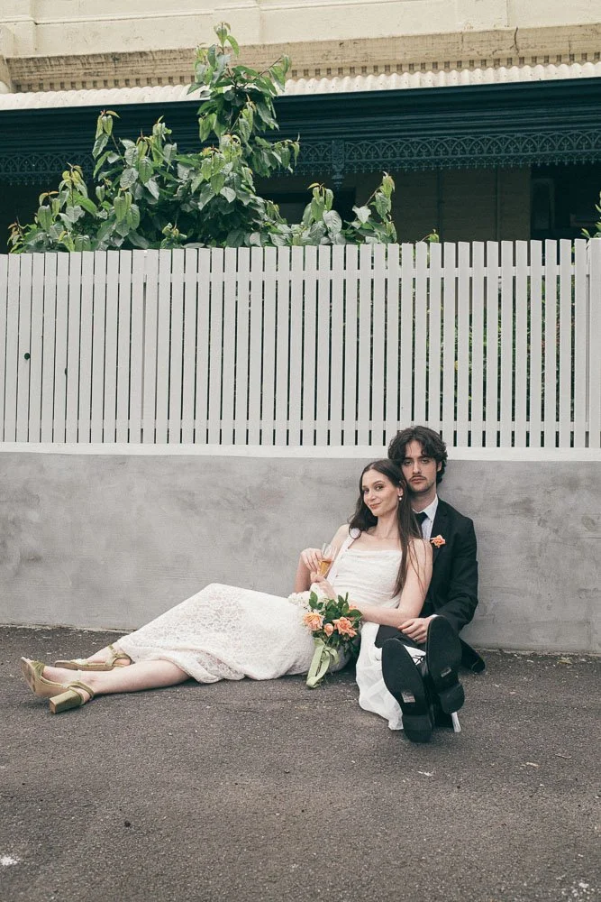 MELBOURNE  EDITORIAL WEDDING PHOTOGRAPHY, BRIDE AND GROOM POSING SITTING ON THE FLOOR AGAINST A GREY WALL AN WHITE FENCE ON THE FLOOR