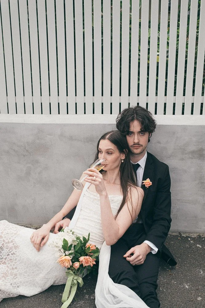 MELBOURNE  EDITORIAL WEDDING PHOTOGRAPHY, BRIDE AND GROOM POSING SITTING ON THE FLOOR AGAINST A GREY WALL AN WHITE FENCE ON THE FLOOR SIPPING CHAMPAGNE