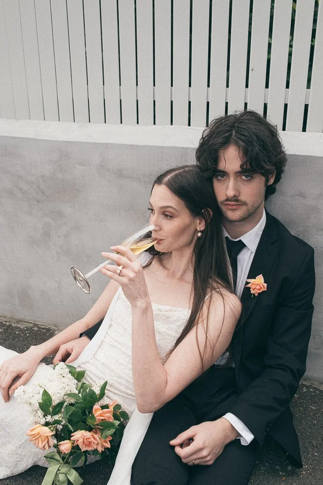 MELBOURNE  EDITORIAL WEDDING PHOTOGRAPHY, BRIDE AND GROOM POSING SITTING ON THE FLOOR AGAINST A GREY WALL AN WHITE FENCE ON THE FLOOR SIPPING CHAMPAGNE