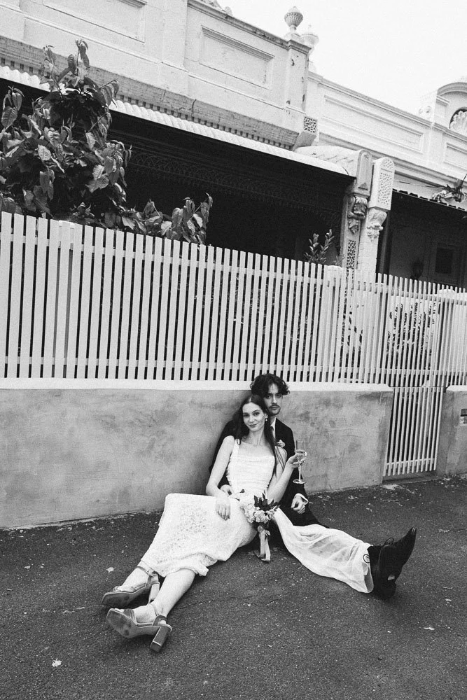 MELBOURNE  EDITORIAL WEDDING PHOTOGRAPHY, BRIDE AND GROOM POSING SITTING ON THE FLOOR AGAINST A GREY WALL AN WHITE FENCE ON THE FLOOR SIPPING CHAMPAGNE