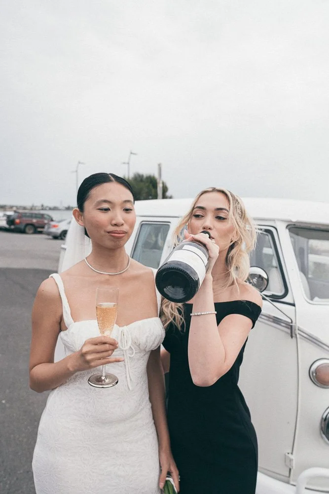 Two women in elegant dresses, one in white and the other in black, standing outdoors near a vintage white van on a cloudy day. One woman is holding a glass of champagne, and the other is drinking from a large bottle.