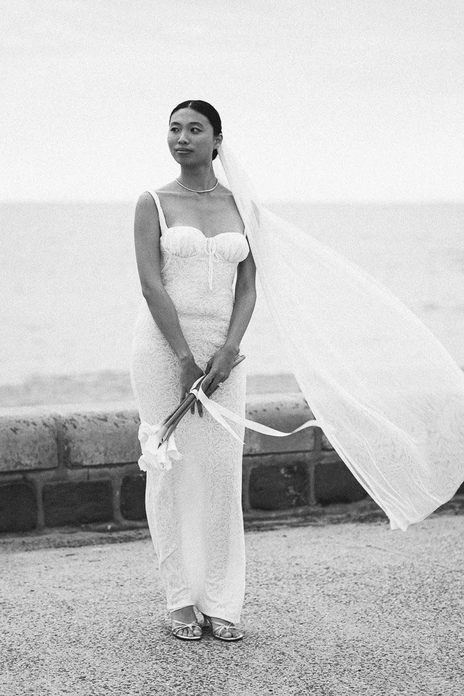 Black and white photo of a woman in a wedding dress holding a bouquet, standing on a sidewalk near a body of water.