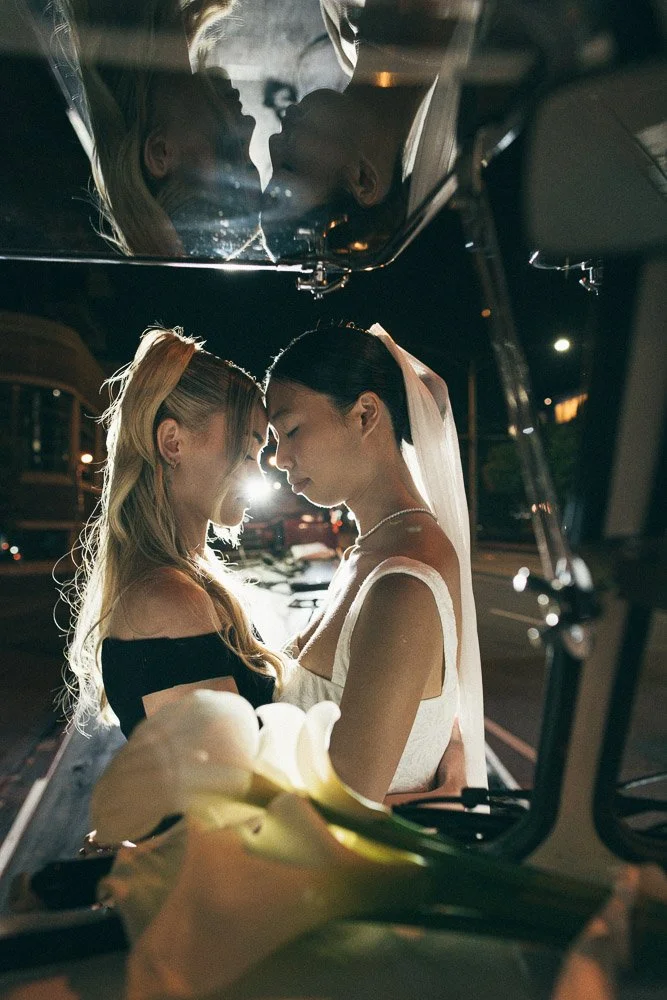 A newlywed couple sharing a kiss at night, seen through the open trunk of a car, with a reflection of their faces on the trunk lid.