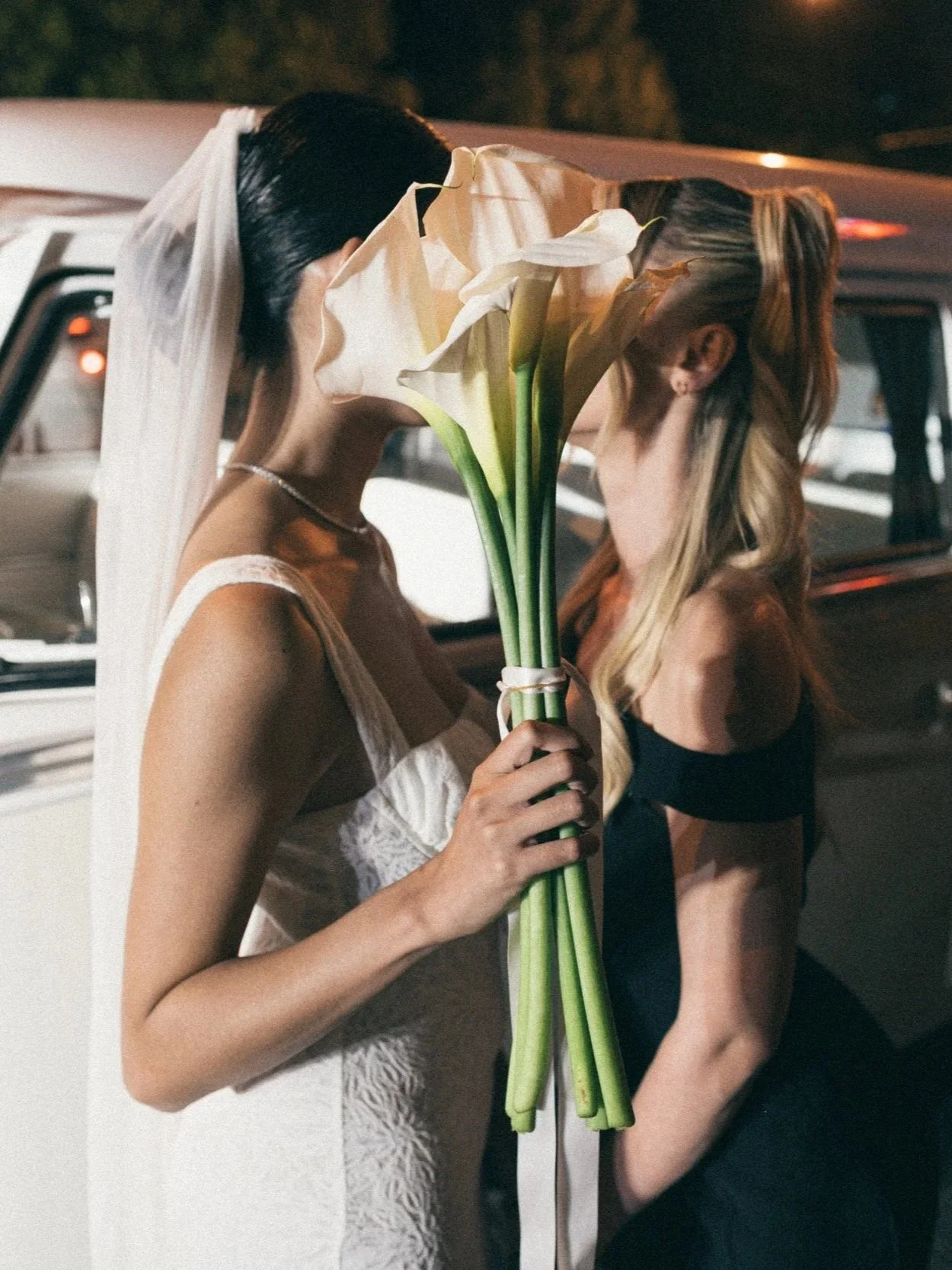 Two women kissing behind a large bouquet of white calla lilies, with one woman in a wedding dress and veil.