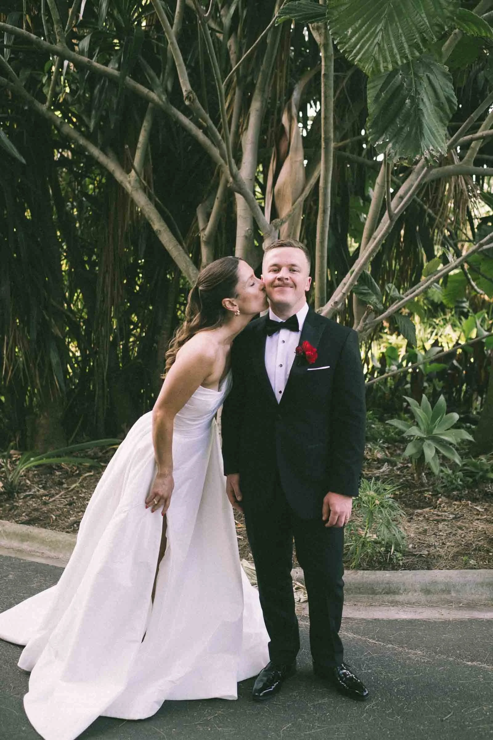 MELBOURNE BOTANICAL GARDEN WEDDING, BRIDE AND GROOM POSING ON THE GARDEN LAWNS, BRIDE KISSING THE GROOM ON THE CHEEKS