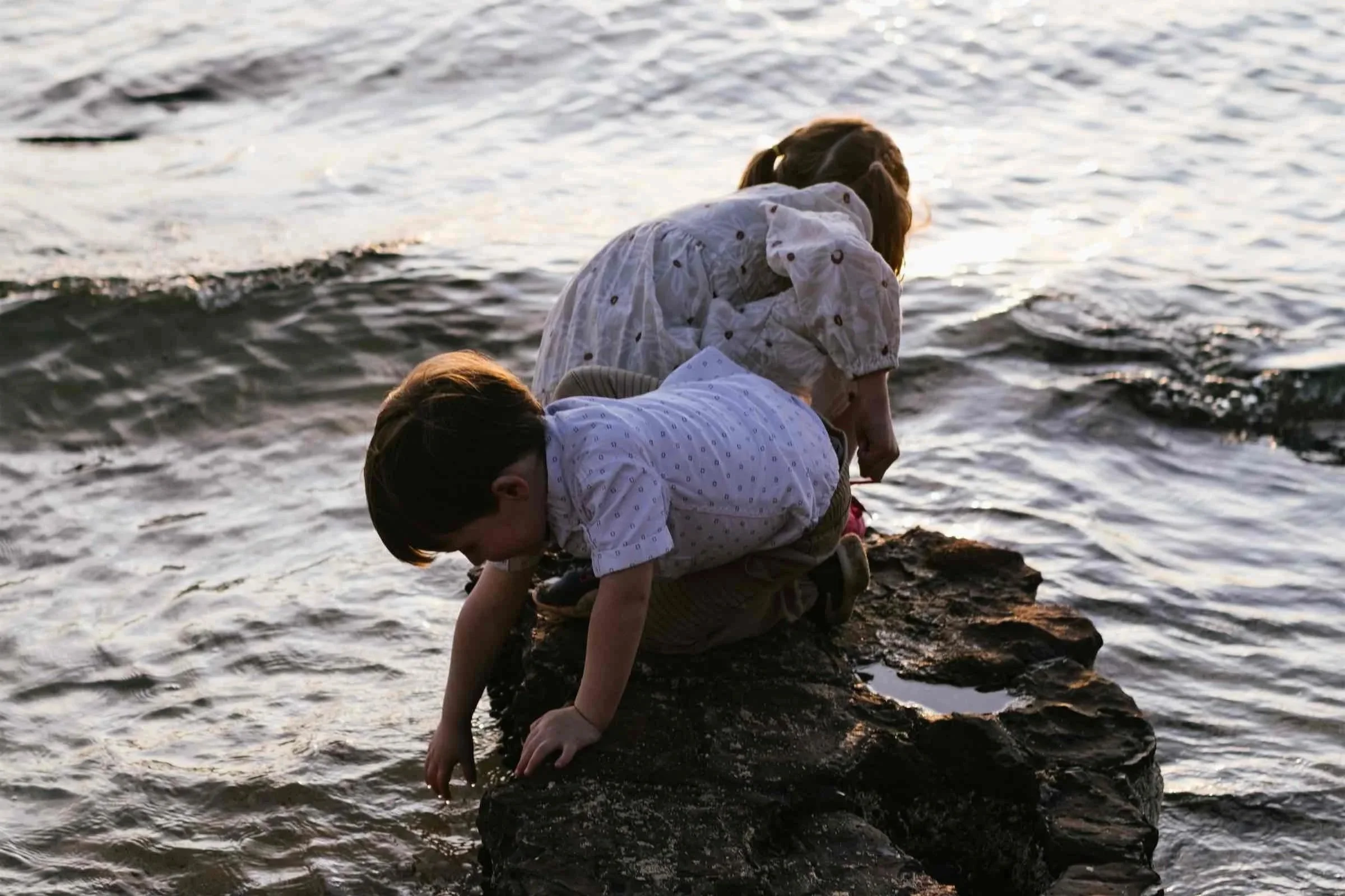 Two children, one wearing a white shirt and one wearing a light-colored dress, are playing near the water on a rocky shore during sunset, with the water shimmering in the background.