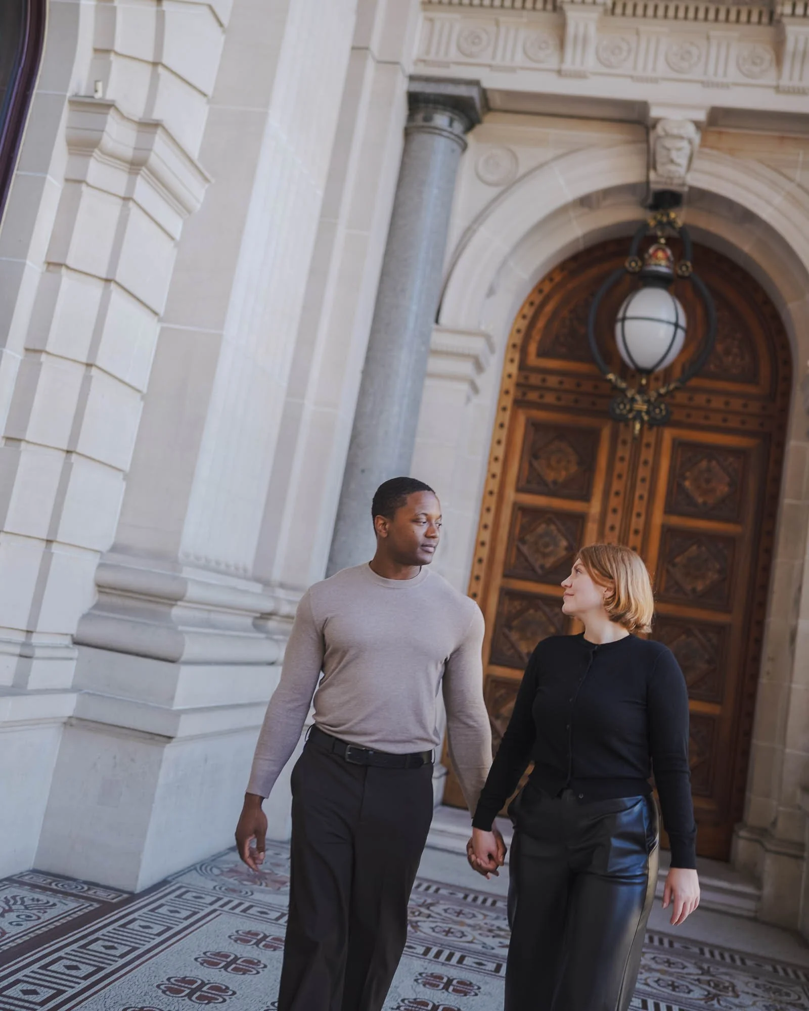A man and woman holding hands and walking together outside a building with ornate wooden doors, decorative stone columns, and patterned tile flooring.