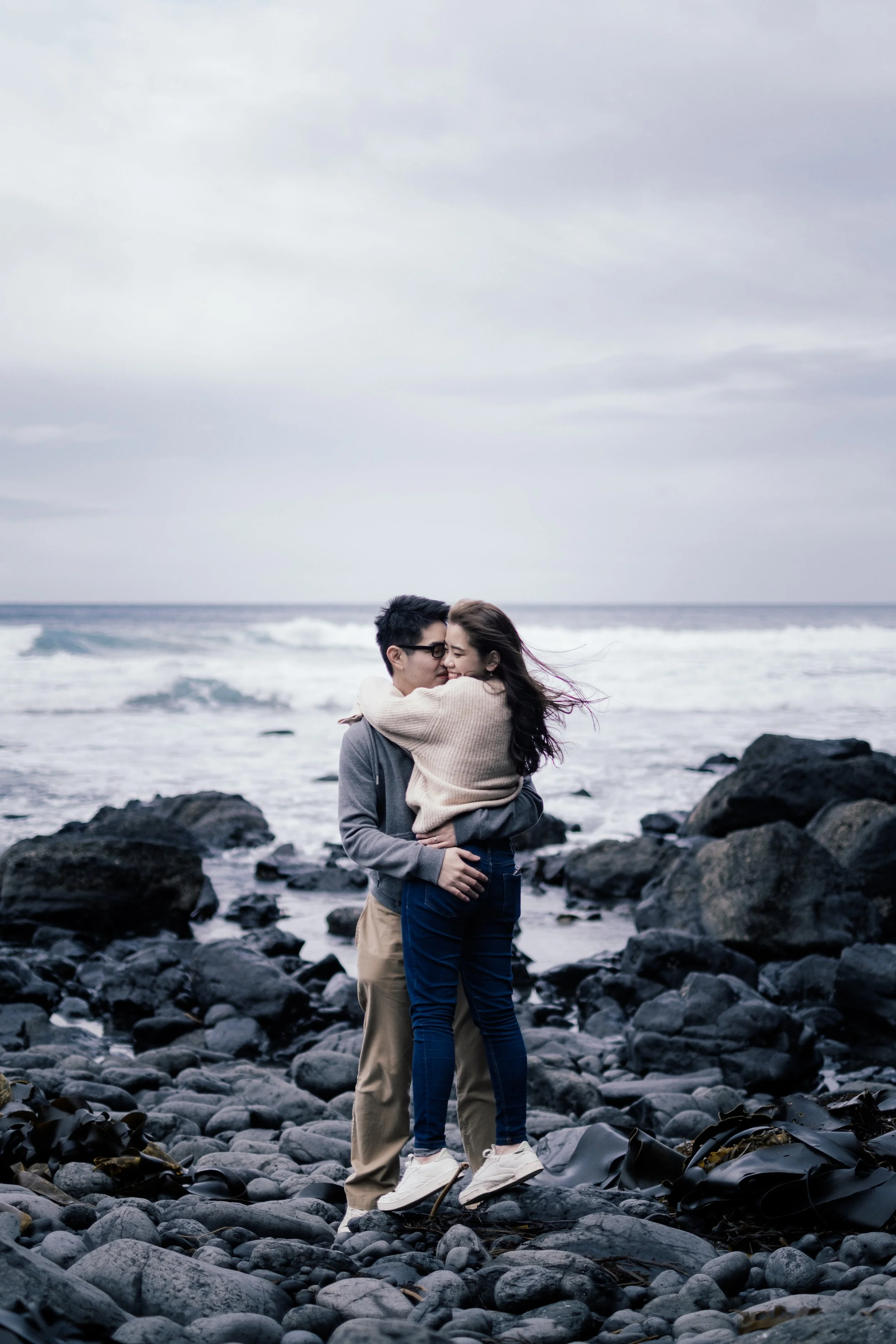 A couple embracing on a rocky beach with ocean waves and an overcast sky in the background.