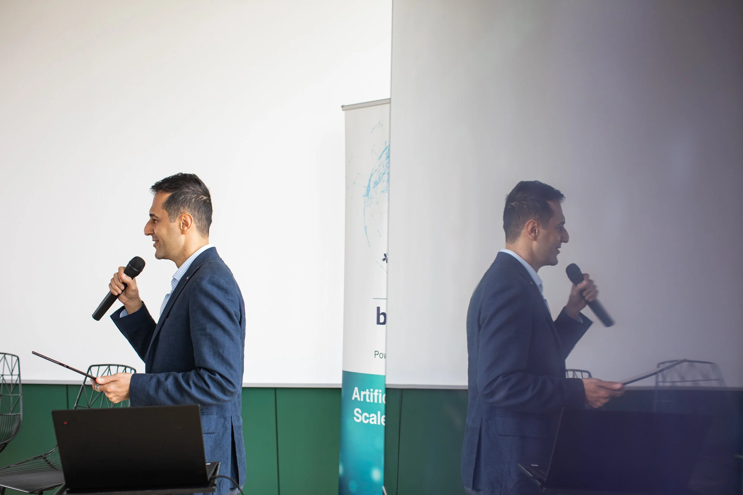 Two men in business suits are speaking into microphones during a presentation or conference. The image is split with one side showing a bright background and the other a reflective surface showing the men turned away. Behind them, there is a banner w