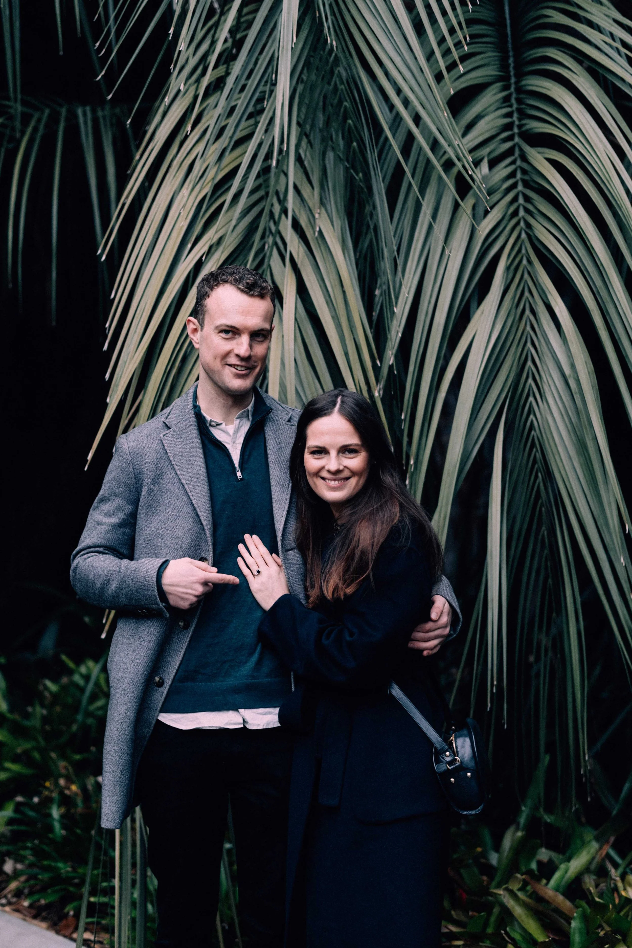 Two smiling people, a man and a woman, standing close together outdoors in front of large green tropical leaves.