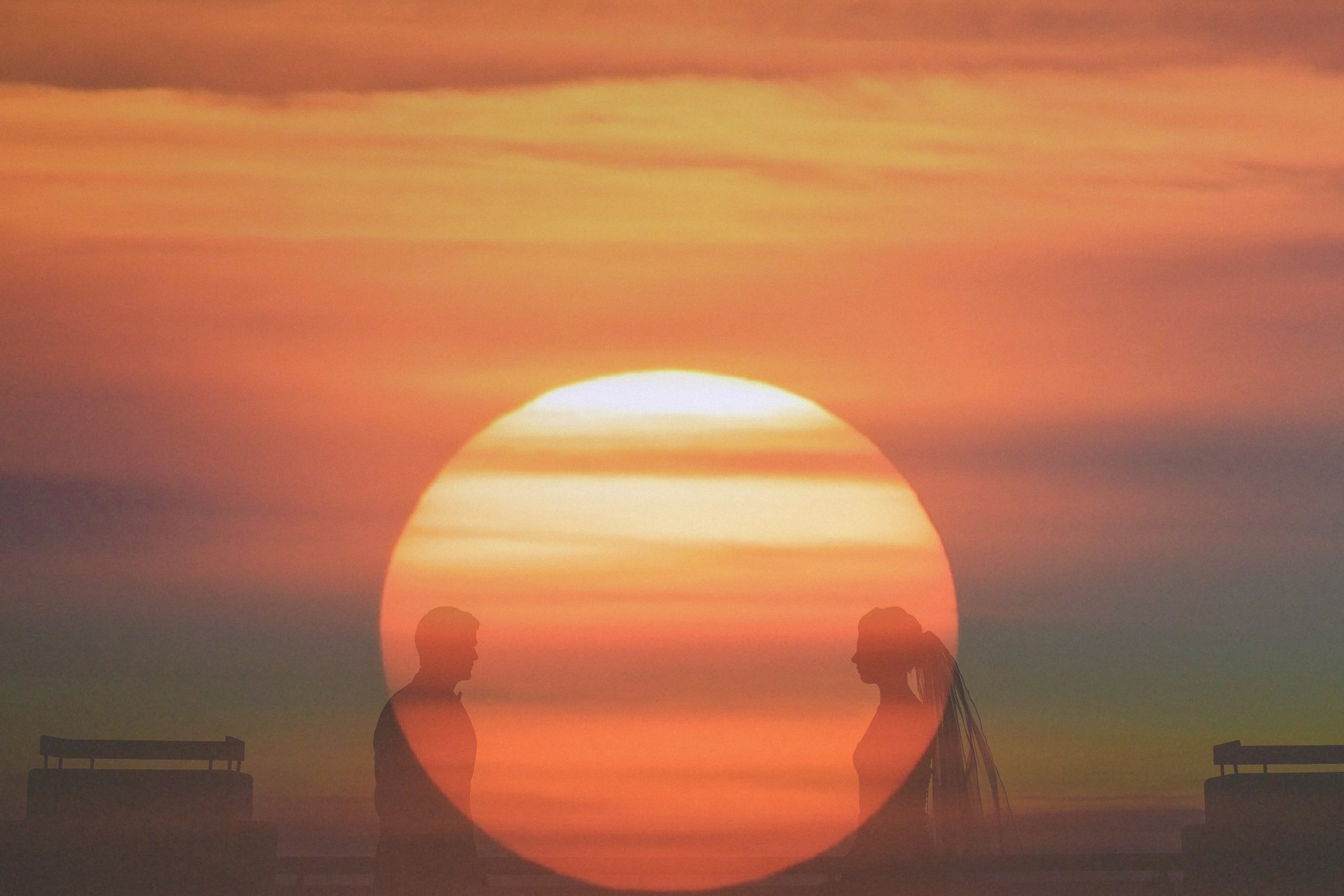 Silhouettes of a man and woman facing each other on a beach during sunset, with the large sun setting on the horizon.