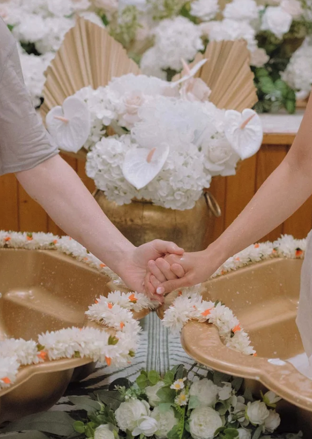 Two people holding hands over a decorated table, with floral arrangements and a gold-colored bowl filled with white flowers in the background, during a wedding or similar ceremony.