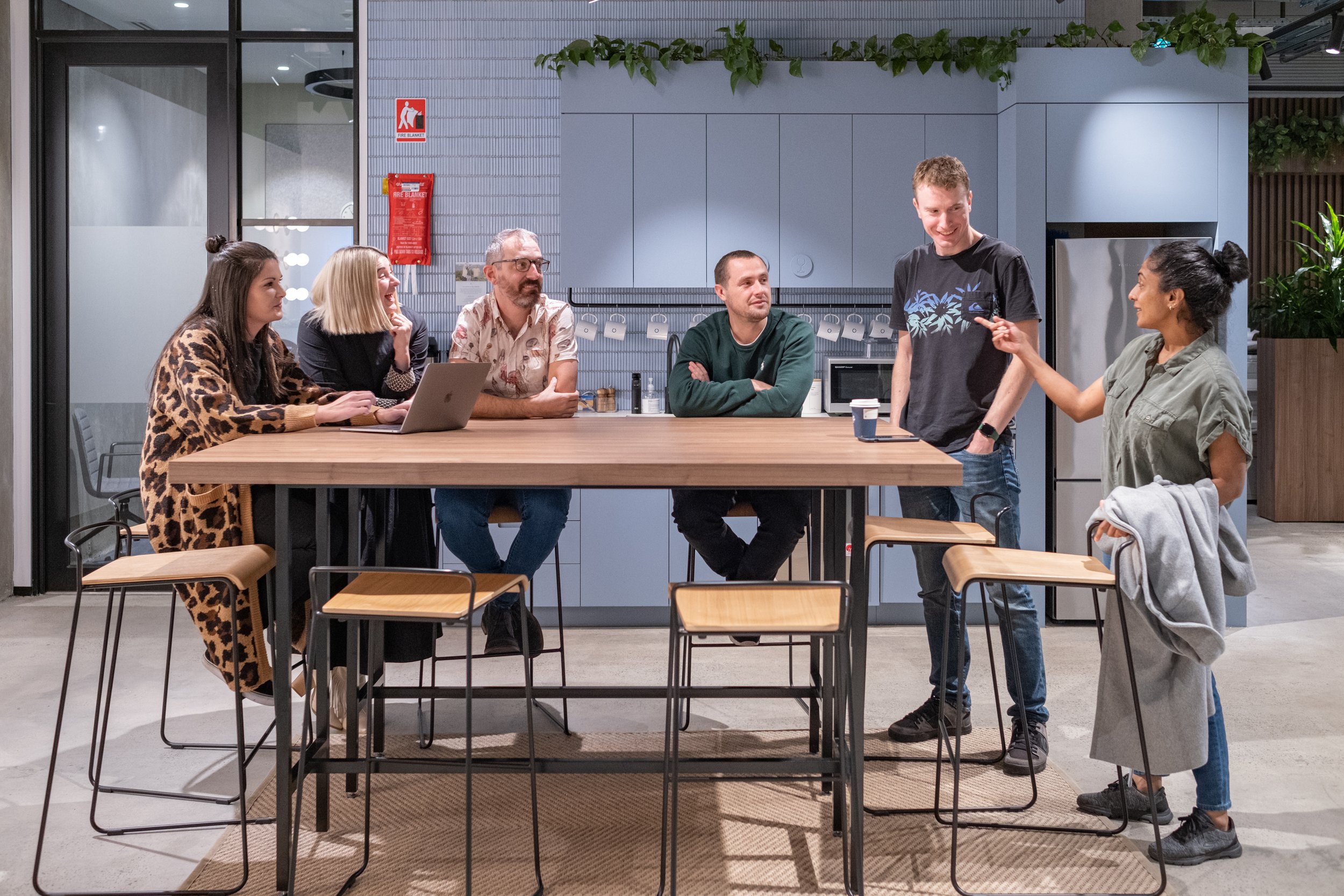 Group of six people gathered around a kitchen island having a conversation, with two women standing and four men sitting, in a modern kitchen setting.