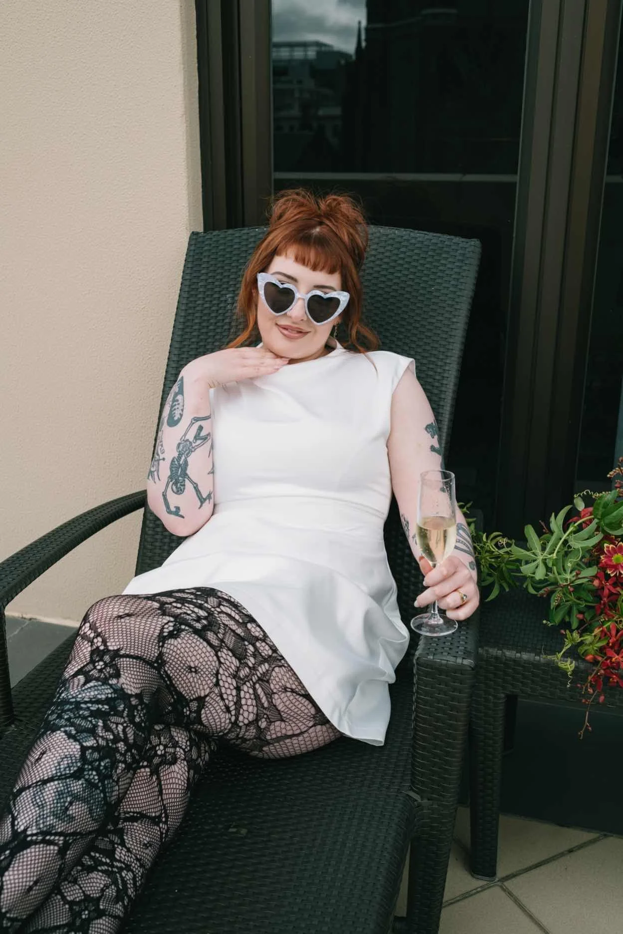 A woman with red hair, tattoos, and heart-shaped sunglasses sits on a black outdoor lounge chair, holding a glass of champagne, wearing a white dress and fishnet stockings with black lace pattern.