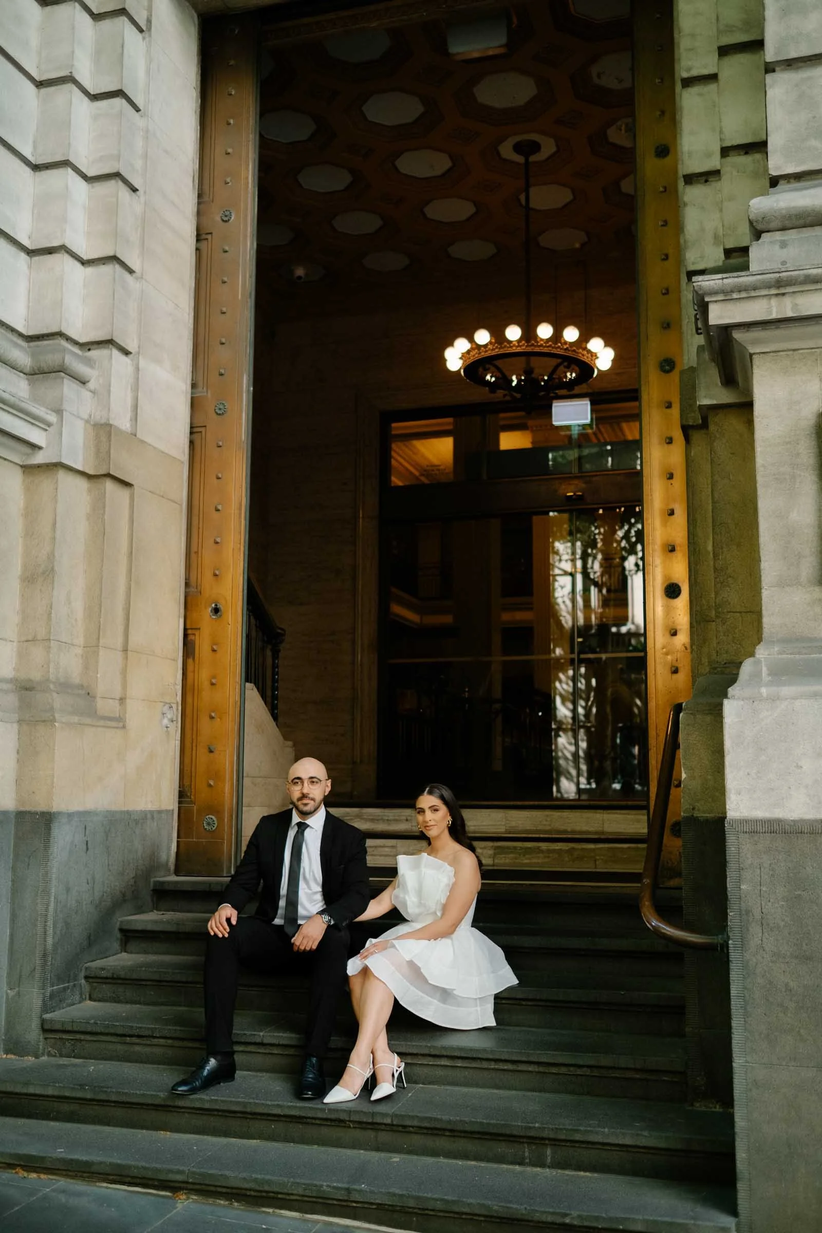 A man and woman sitting on the steps of a building. The man is wearing a black suit with a white shirt and a black tie. The woman is wearing a white dress. The building has stone stairs and tall stone walls with decorative architectural elements, and
