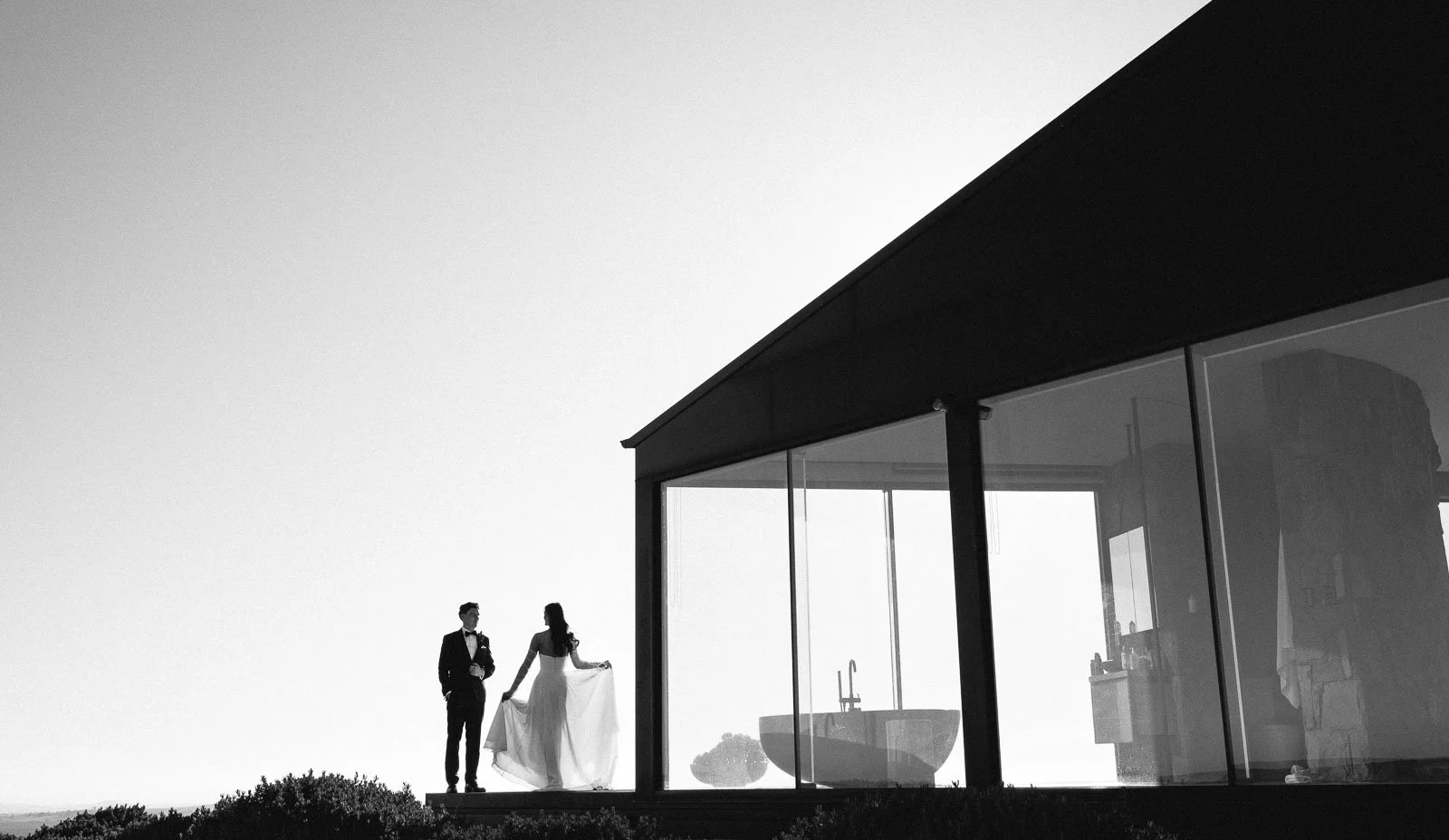 A black and white photo of a wedding couple holding hands outside a modern glass house. The man is in a tuxedo, and the woman is in a wedding dress, holding the dress's train.