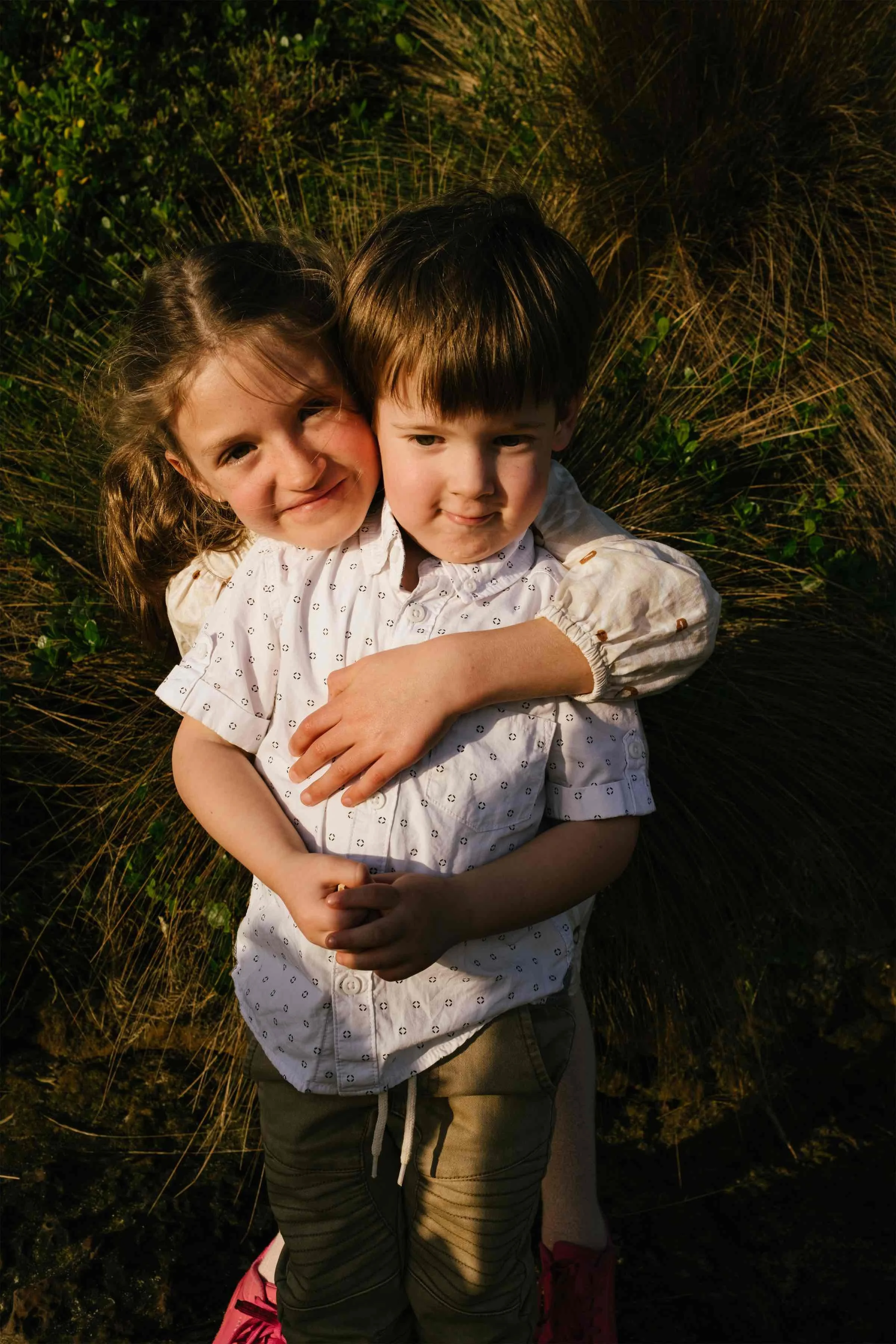 Two children, a girl and a boy, hugging outdoors among tall grass and green plants during sunset.