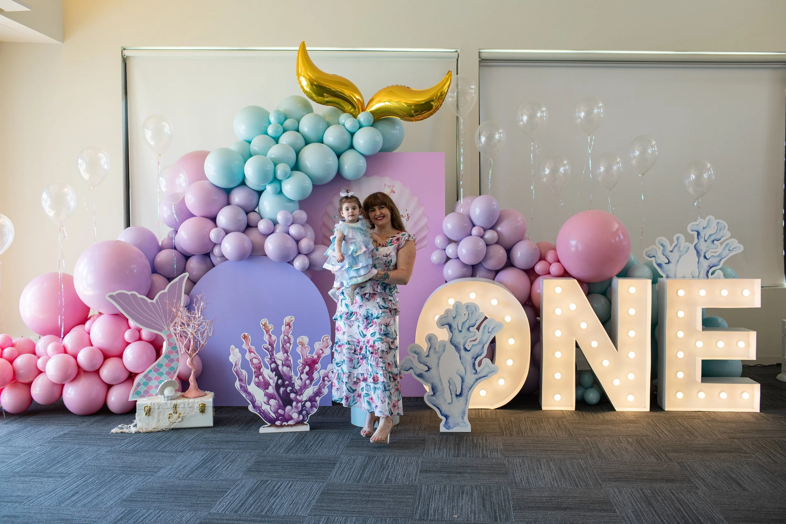 A woman holding a young girl in front of an underwater-themed birthday display with pink, purple, and blue balloons, coral cutouts, and large decorative letter signs.