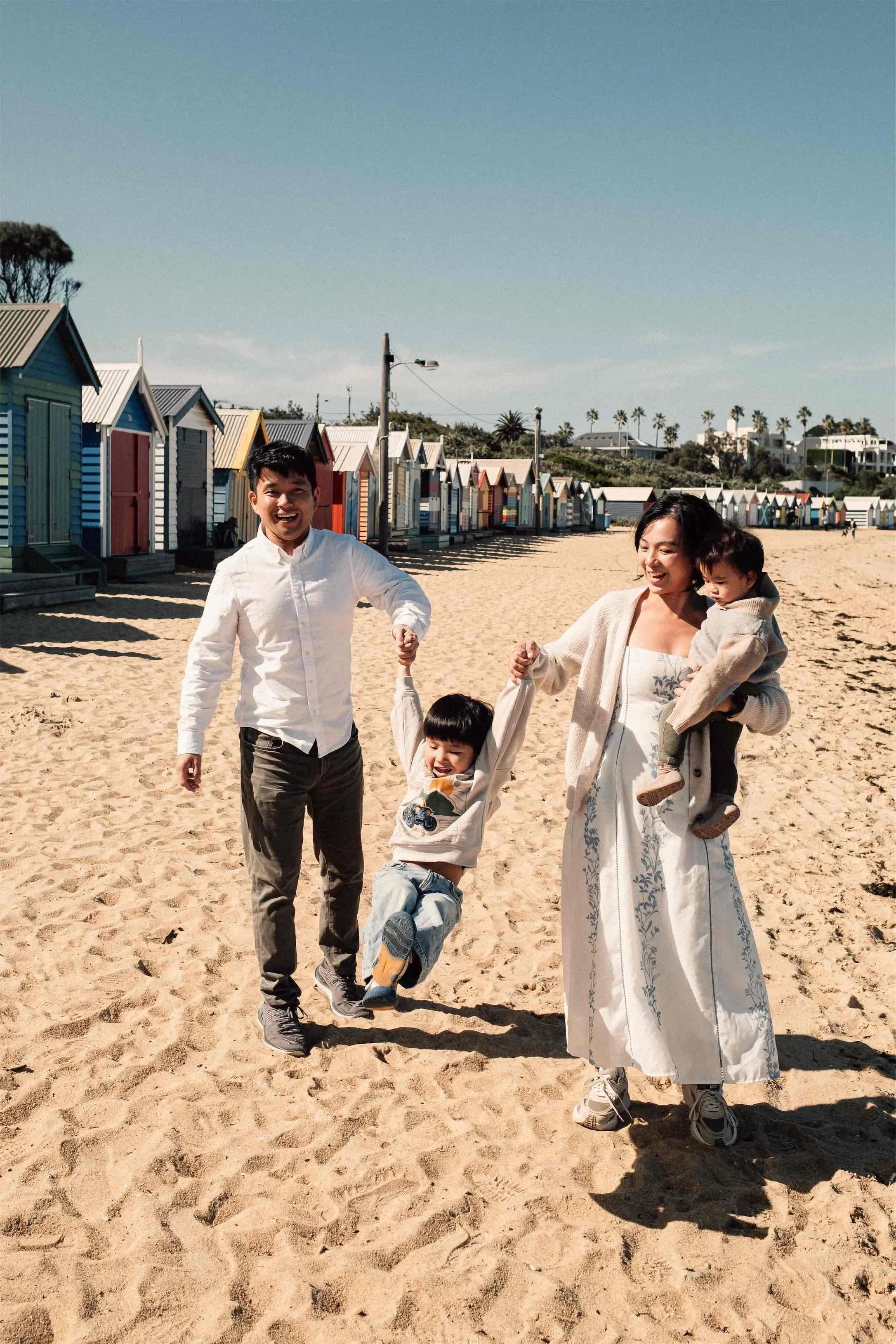 A family of four enjoying a day at the beach, holding hands and smiling. The father is wearing a white shirt and gray pants, the mother is in a long white dress with blue embroidery, and the two children are dressed in casual clothes, one of them bei