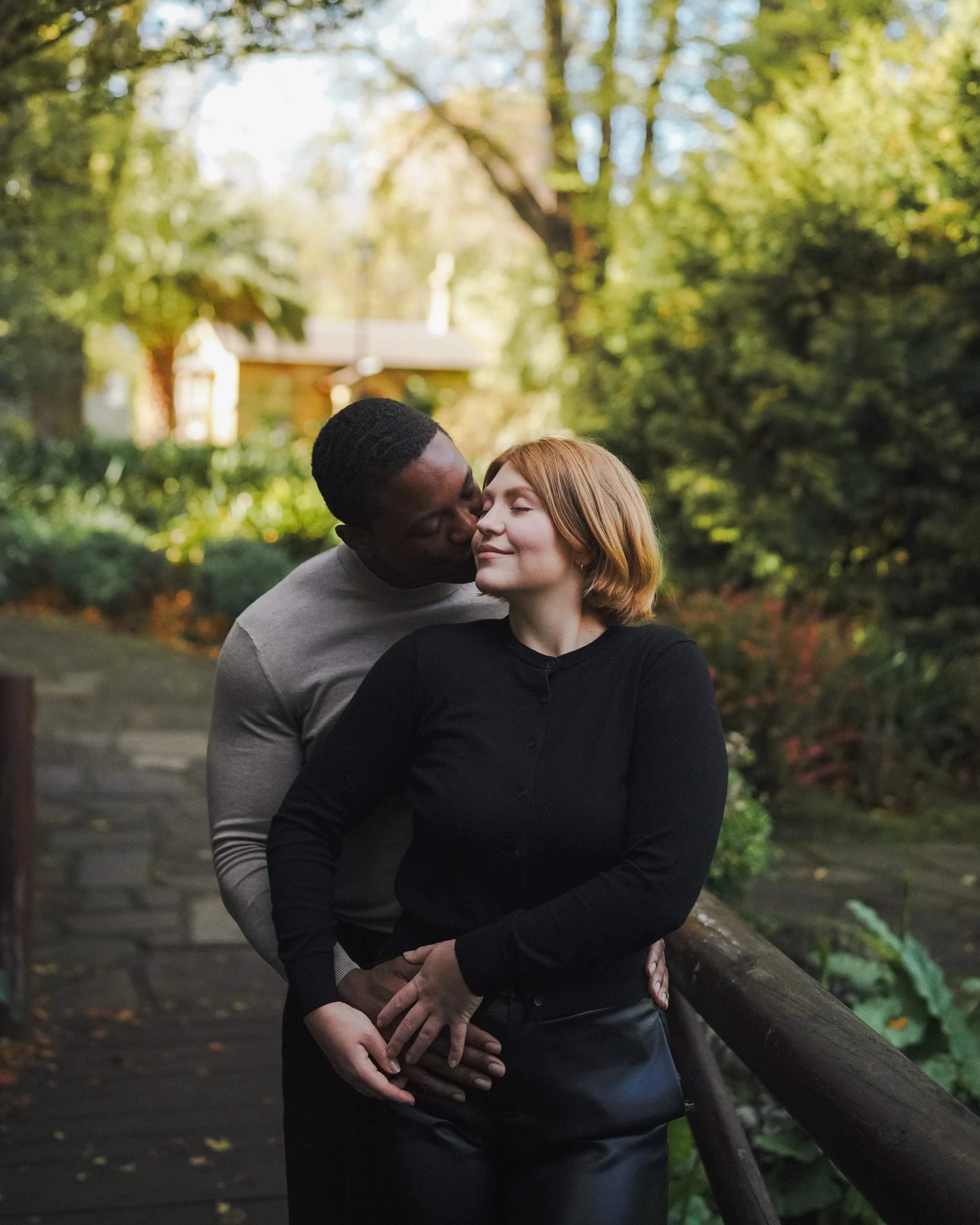 A couple sharing a romantic moment outdoors, with the man kissing the woman's cheek and the woman smiling gently, surrounded by lush green trees.