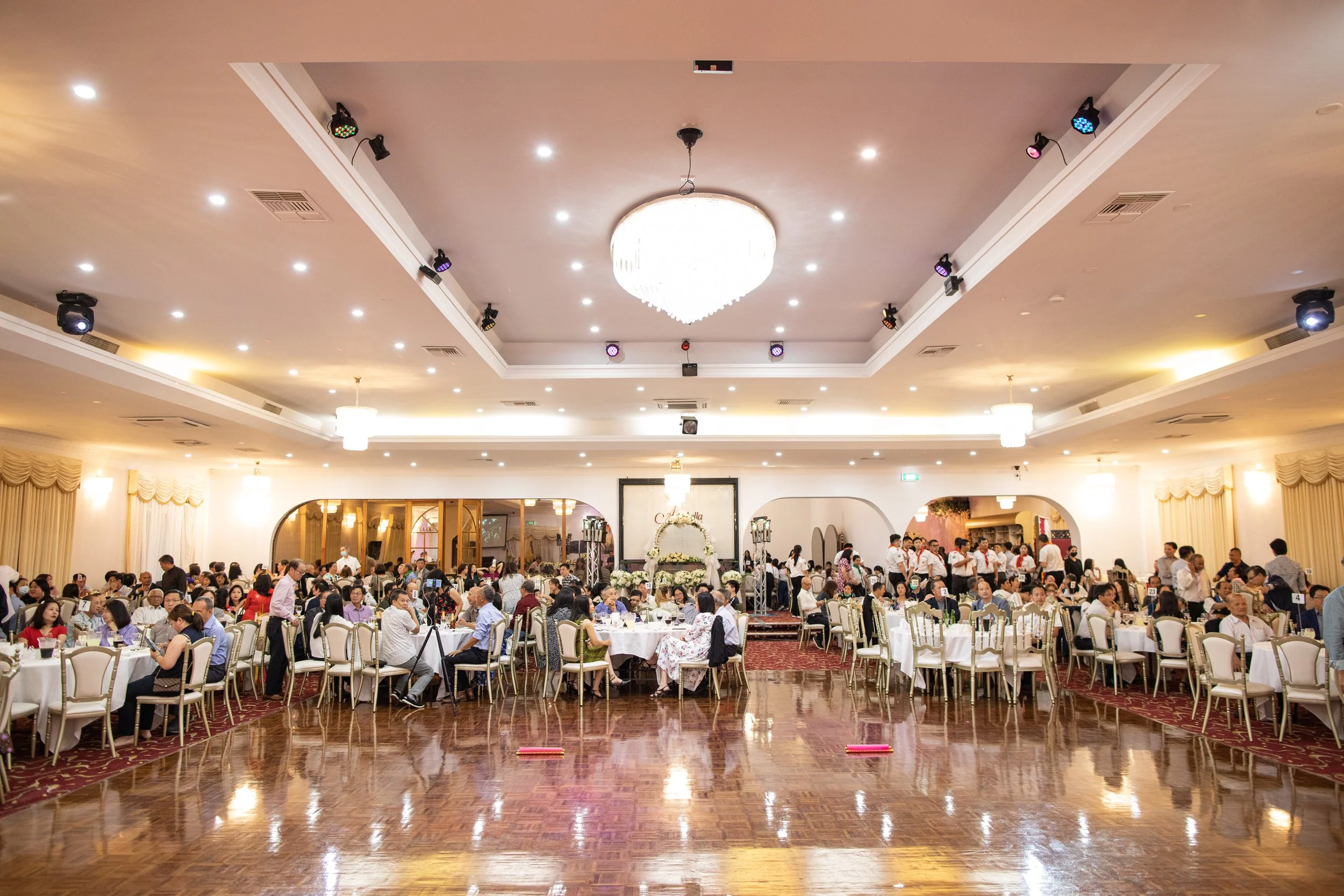 Large banquet hall filled with numerous round tables, each surrounded by chairs and occupied by guests attending an event. The ceiling features a large crystal chandelier and several smaller lights, with stage lighting equipment visible. The area has