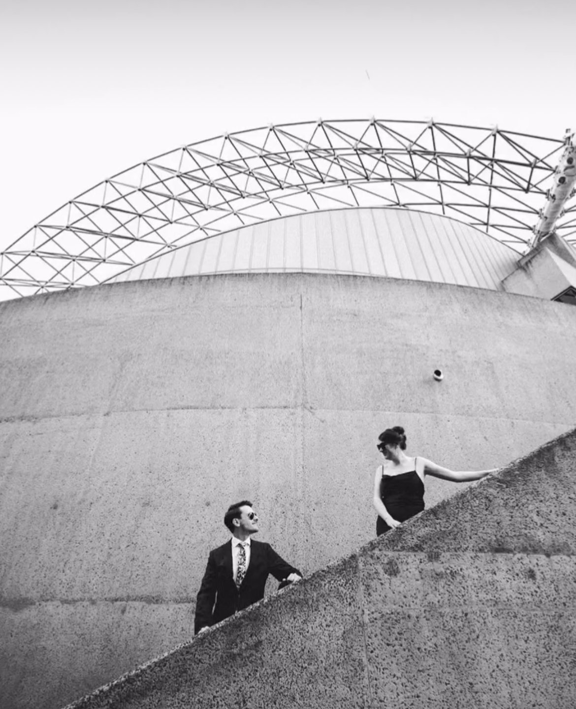 Black and white photo of a man and woman in formal attire ascending a concrete staircase against a modern architectural backdrop.