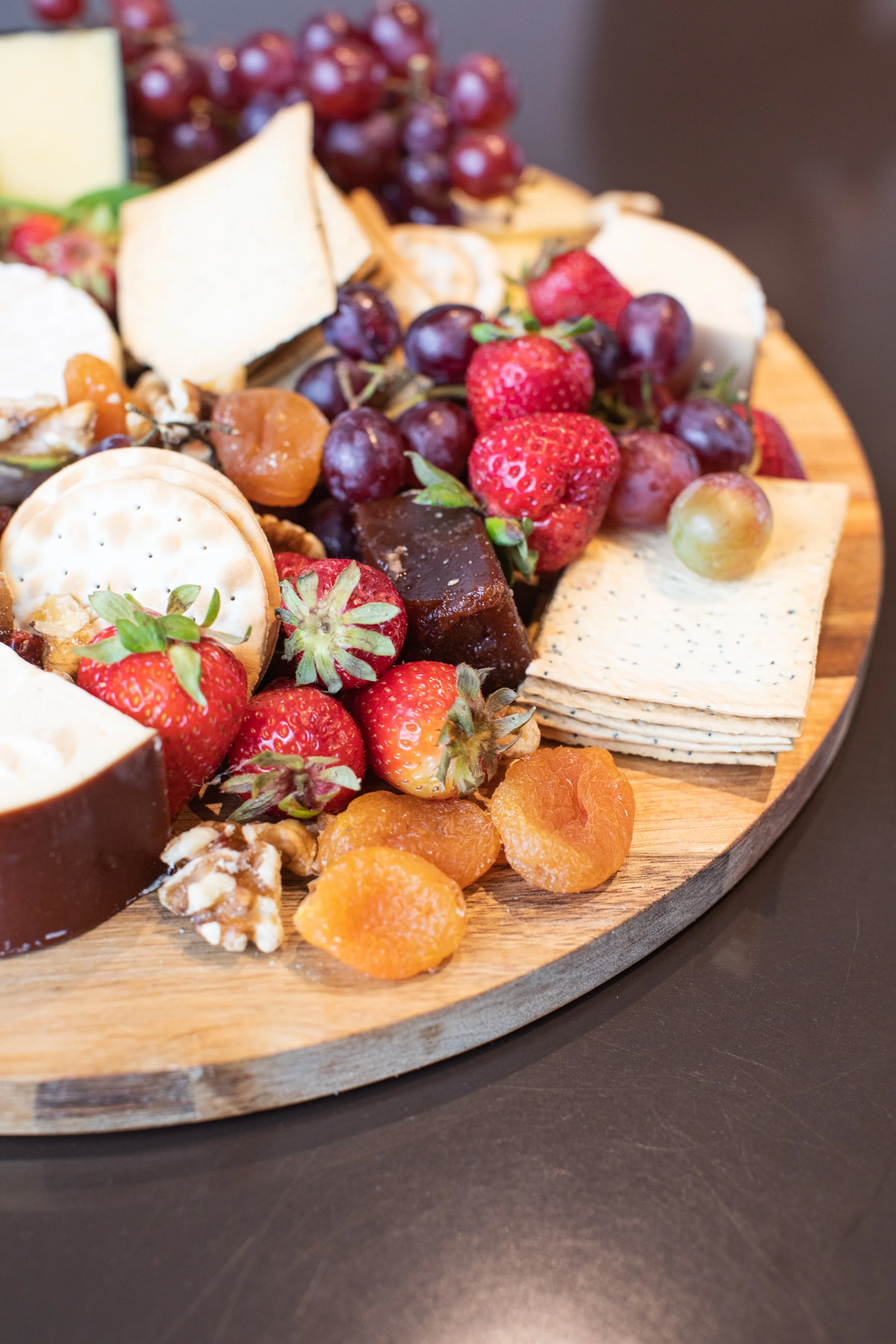 Close-up of a wooden cheese board with grapes, strawberries, crackers, cheese, and dried apricots.