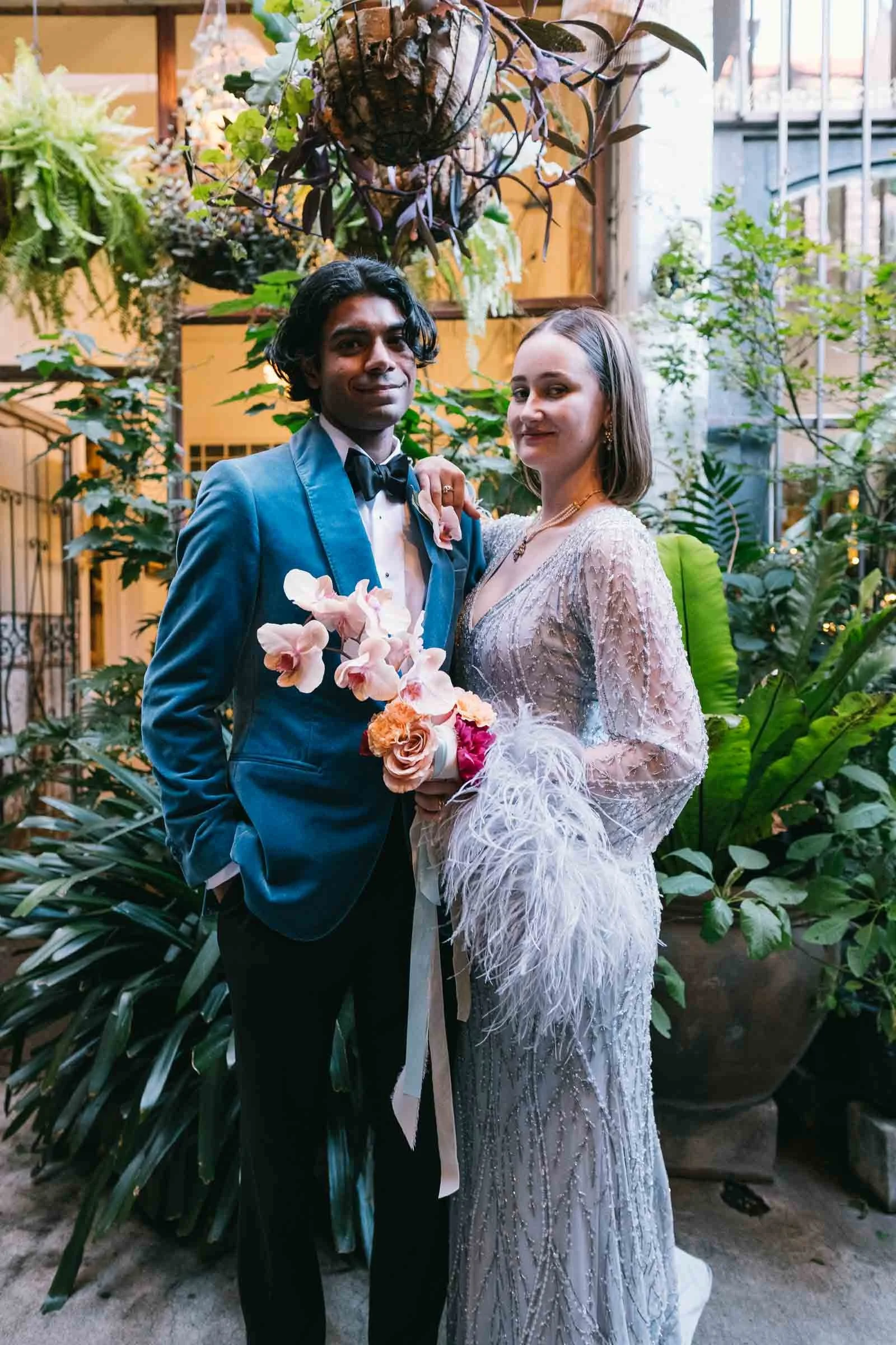 A couple in wedding attire standing in an indoor garden with lush green plants and hanging decorations, smiling at the camera.