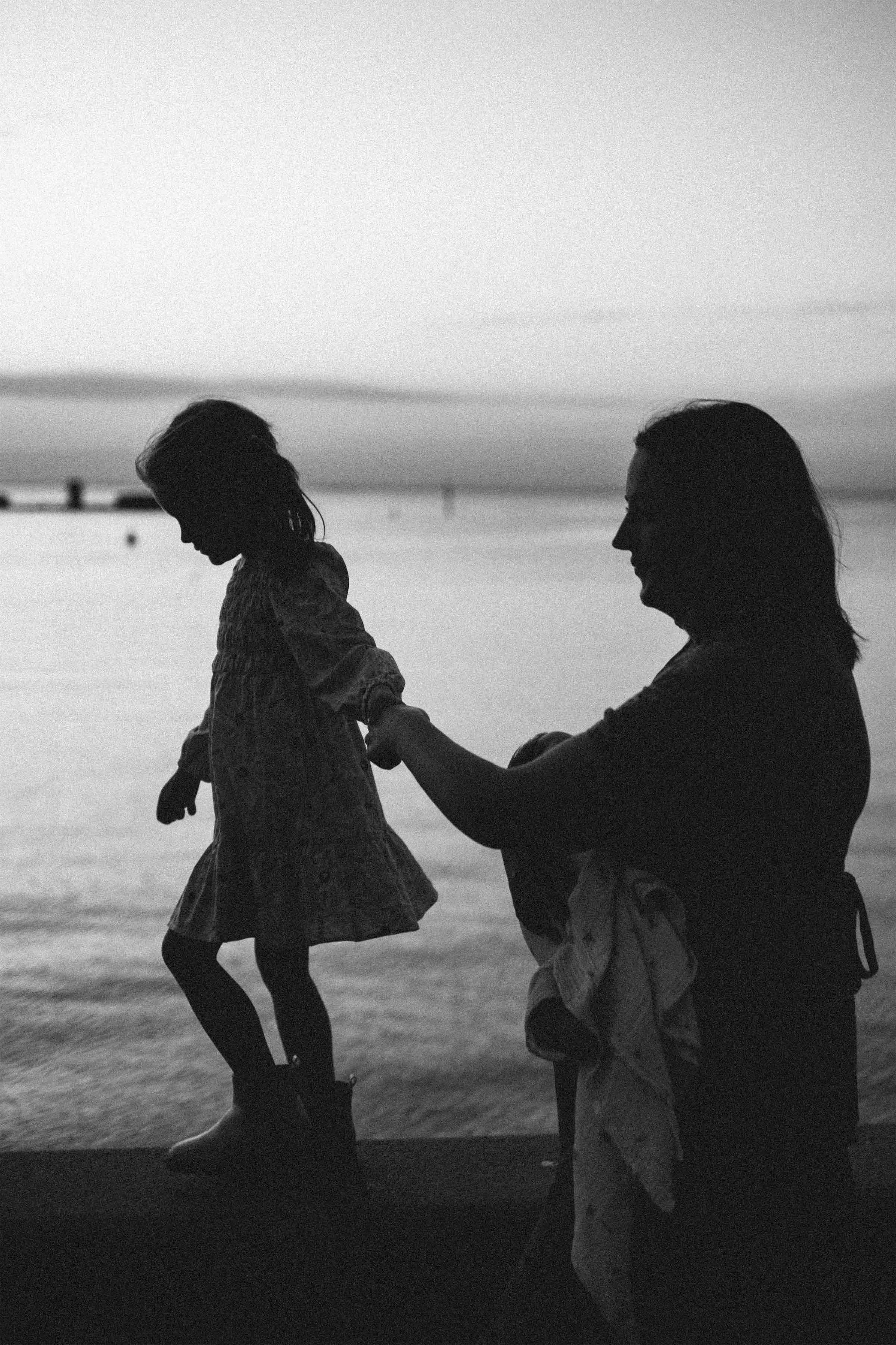 Black and white photo of a woman and a young girl holding hands at the beach, walking along the shoreline at sunset or sunrise.