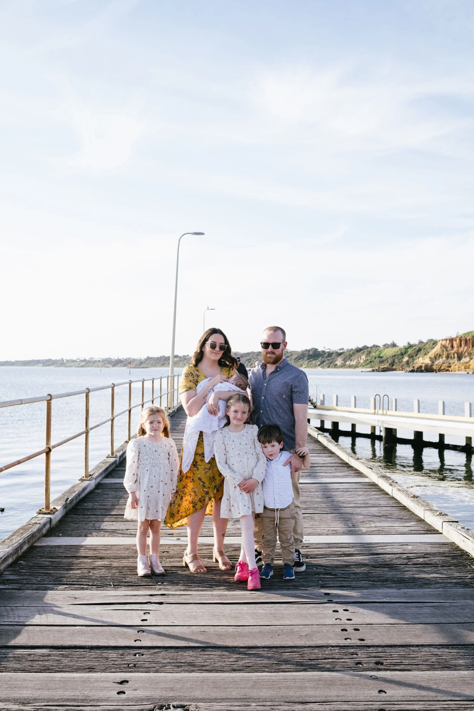 A family of seven standing on a wooden pier over water, with cliffs and a cloudy sky in the background. The mother is holding a baby, and there are five children; three girls in matching cream dresses, a boy in a white shirt and beige pants, and a girl in a mustard yellow dress.