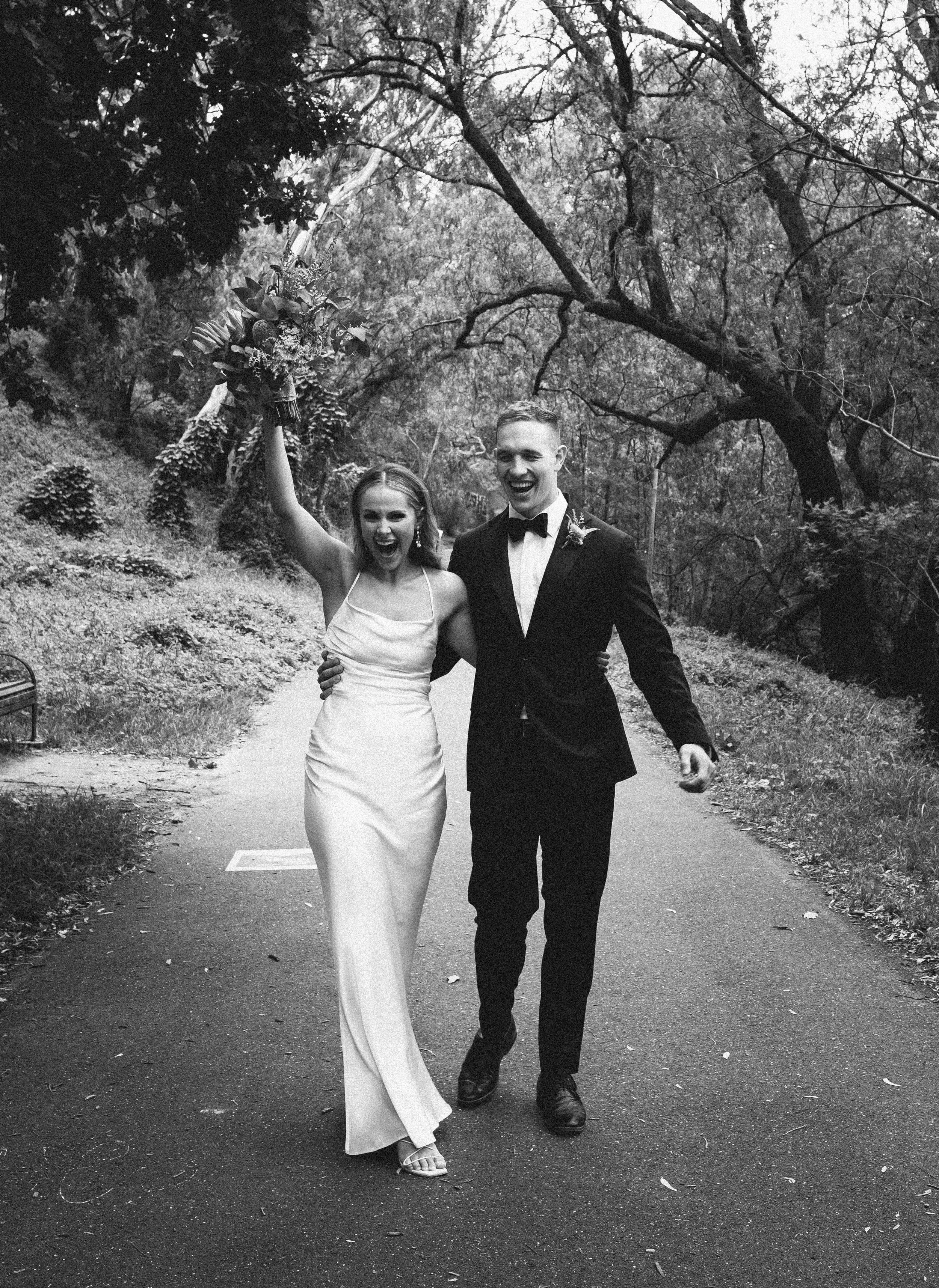 Black and white photo of a bride and groom walking outdoors on a path, with trees in the background. The bride is holding a bouquet up and smiling, and the groom has his arm around her, both looking very happy.