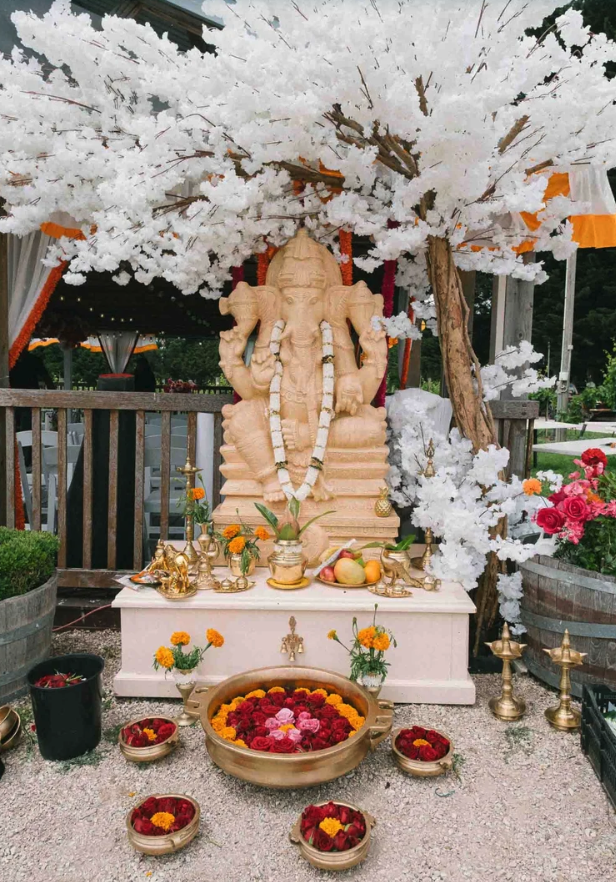 A carved stone Ganesh idol decorated with a flower garland and placed on a white platform, surrounded by offerings including fruits, flowers, and traditional items, under a white artificial cherry blossom tree at a Hindu religious site.