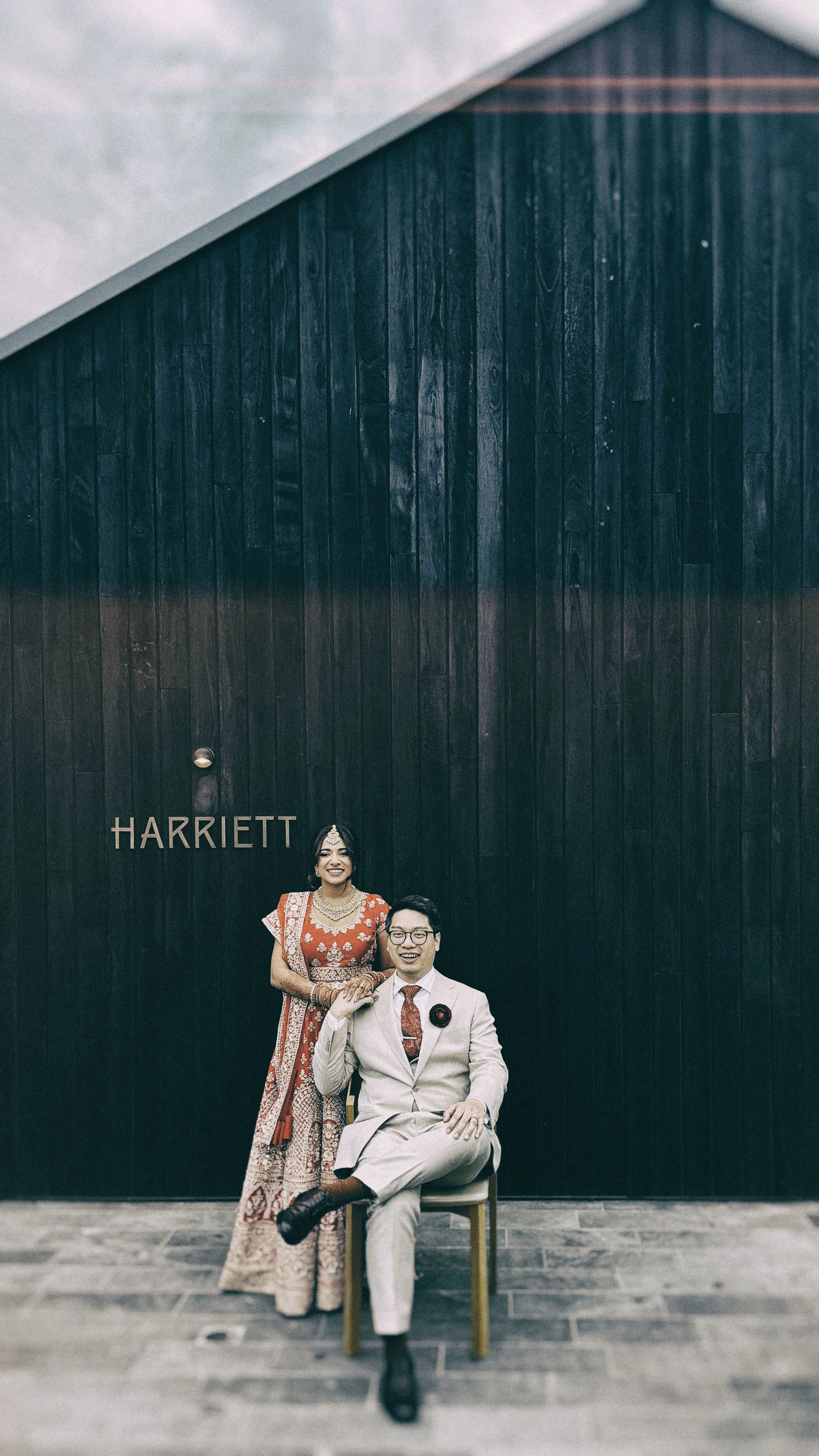 A couple dressed in formal attire posing in front of a dark wooden wall with the word "HARRIETT" written on it. The woman is standing behind the man, both smiling.