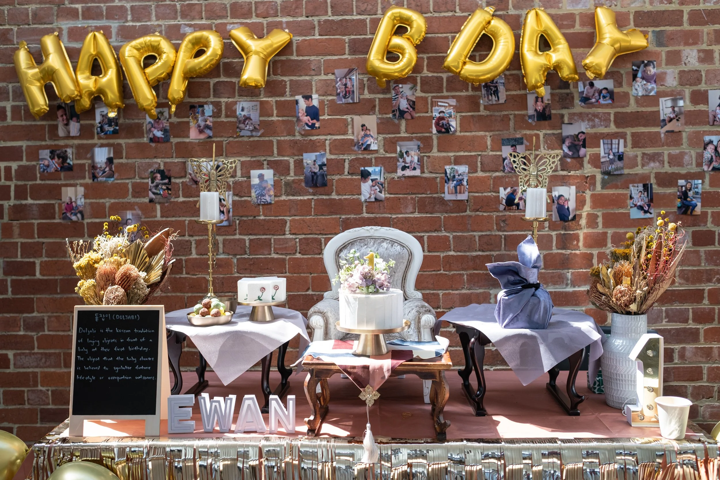 Decorative birthday setup with gold balloons spelling 'HAPPY 6 BABY' hanging against a brick wall. Small photos of children and an adult are pinned on the wall. A table with flowers, candles, and a blackboard sign explaining Doljabi, a Korean traditi