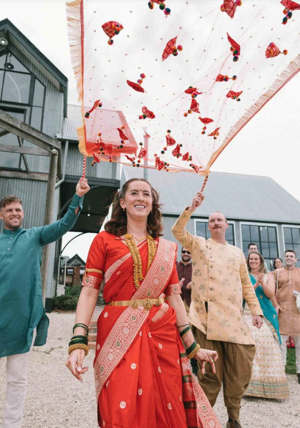 A woman dressed in a red traditional Indian saree with gold jewelry and bangles, holding a decorated umbrella, accompanied by men and women in traditional Indian clothing at an outdoor celebration or wedding. They are standing on gravel with a barn o
