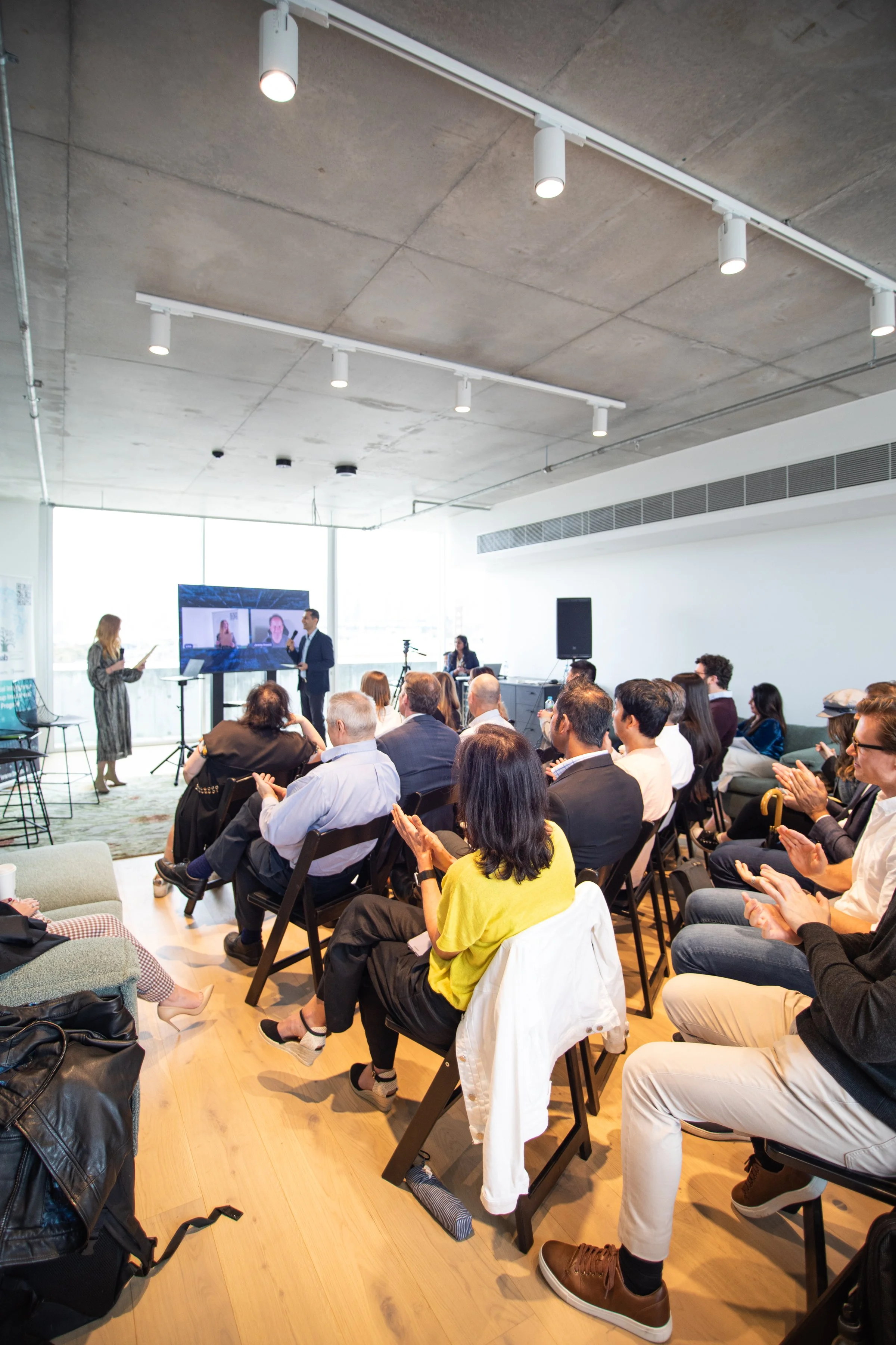 A group of people attending a conference or presentation in a modern, spacious room with large windows. Two presenters are standing near a large screen displaying a person's face. The audience is sitting in chairs, clapping, and some are using tablet