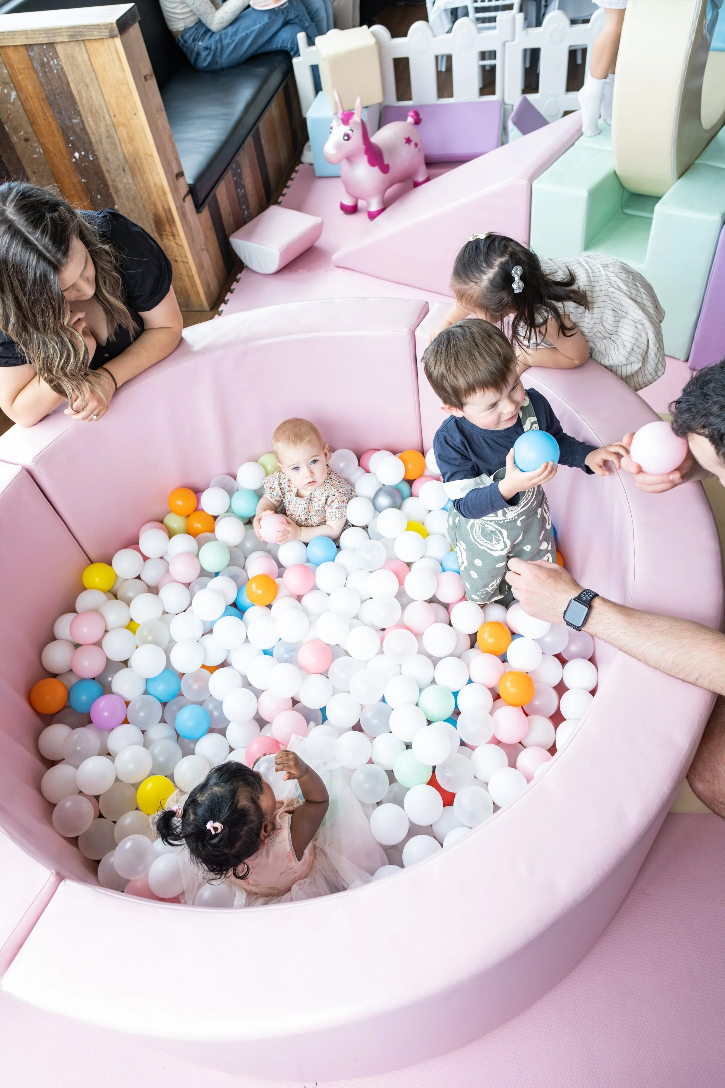 Children playing in a pink ball pit with colorful plastic balls, a pink unicorn ride in the background, and adults supervising.