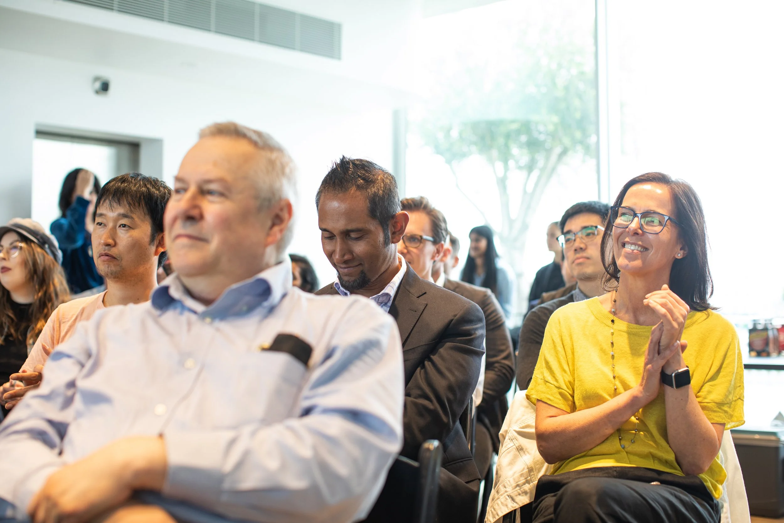 Group of diverse people attending a conference or seminar, sitting in chairs, some clapping, in a well-lit modern room with large windows and green trees outside.