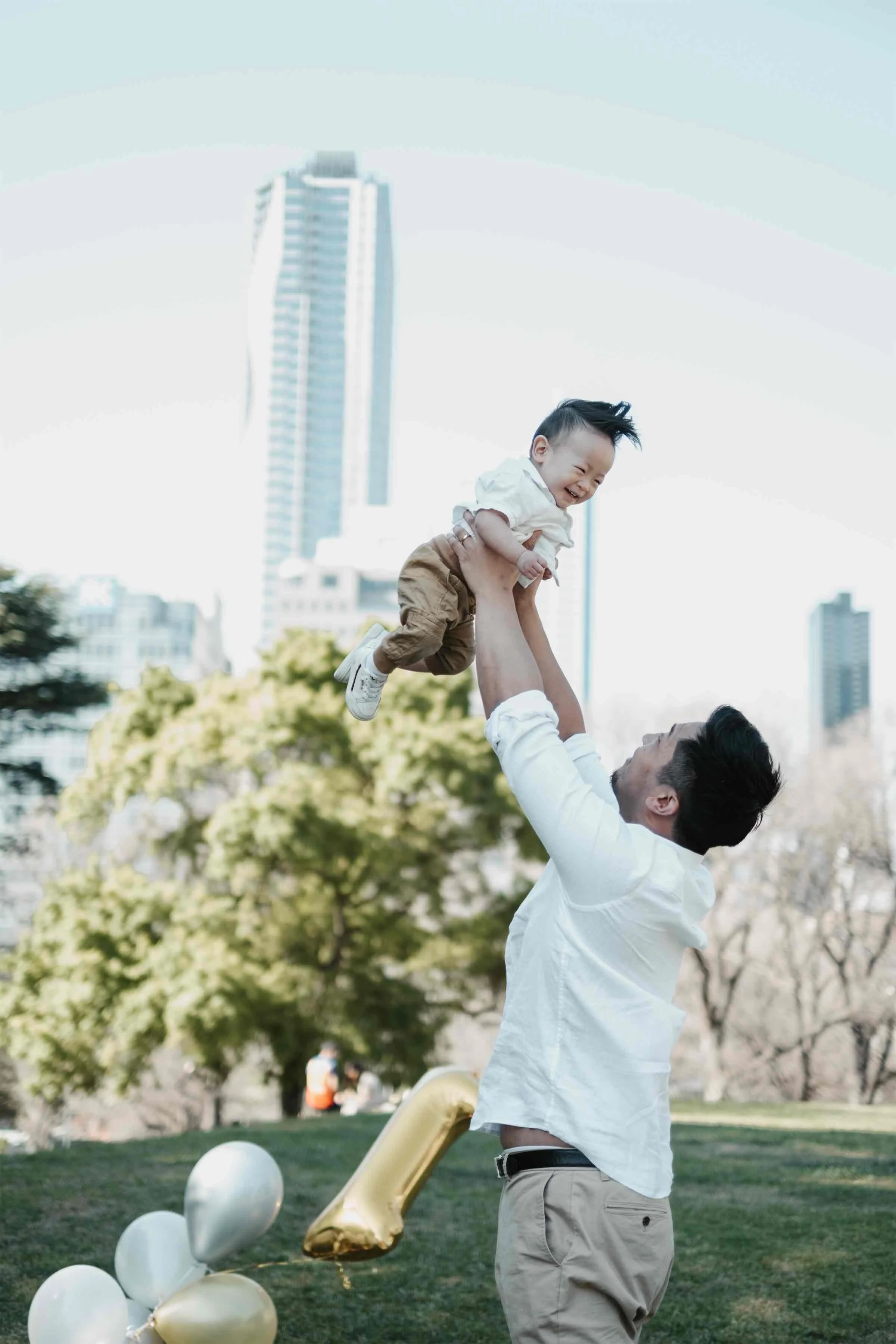 A man lifting a smiling young boy in the air outdoors, with balloons and a cityscape with tall buildings in the background.
