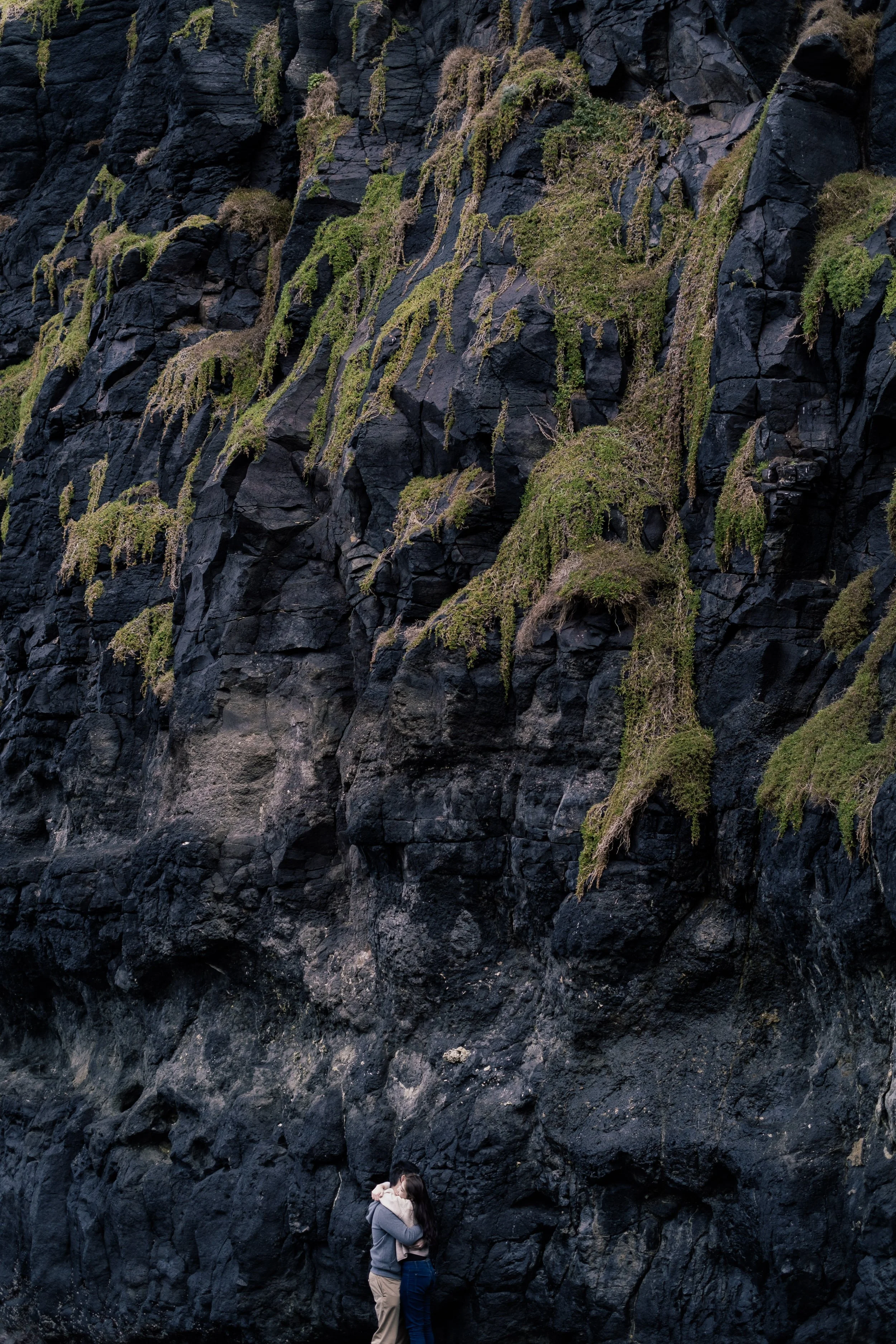 A couple hugging in front of a tall, dark rocky cliff face with sparse green vegetation.