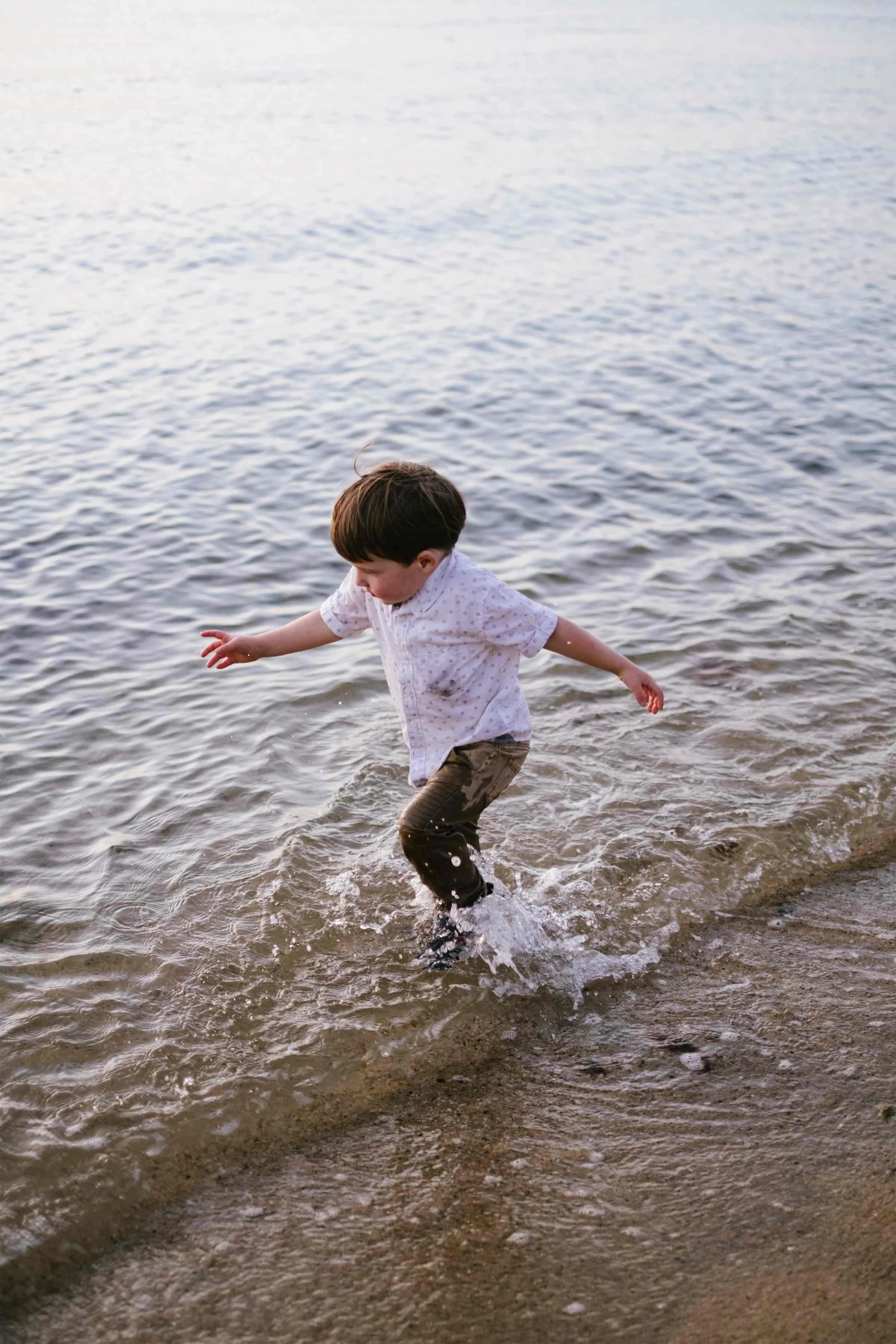 A young boy with brown hair, wearing a white shirt and pants, playing in shallow water at the edge of a lake or ocean.