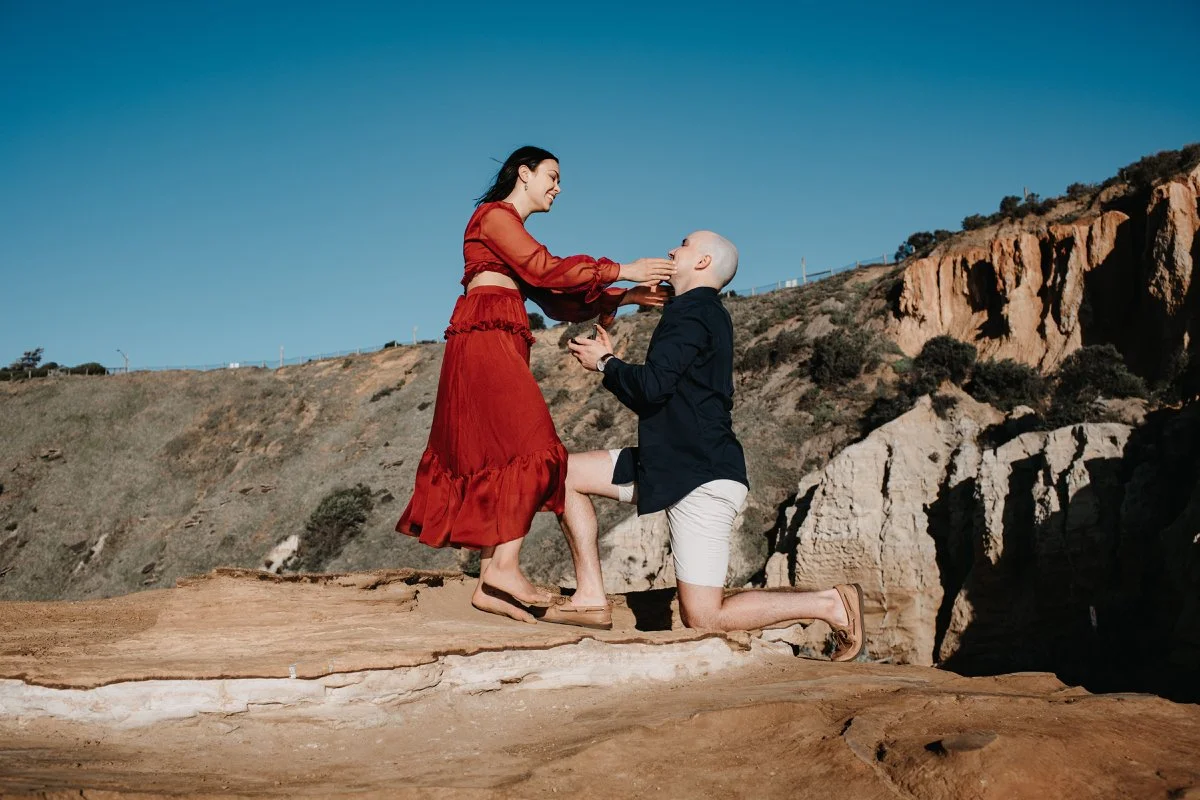 A man kneeling on one knee on a rocky surface, holding a woman's hands as he proposes, with a woman wearing a red dress smiling and reaching out to him, in an outdoor desert landscape.