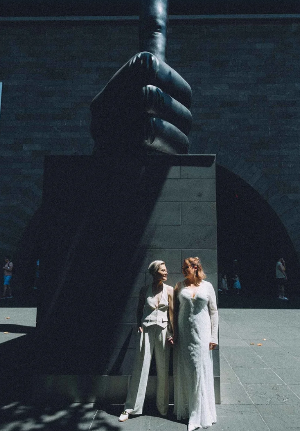 Two women stand holding hands in front of a tall, dark sculpture of a hand gripping a pole, with a brick building in the background.