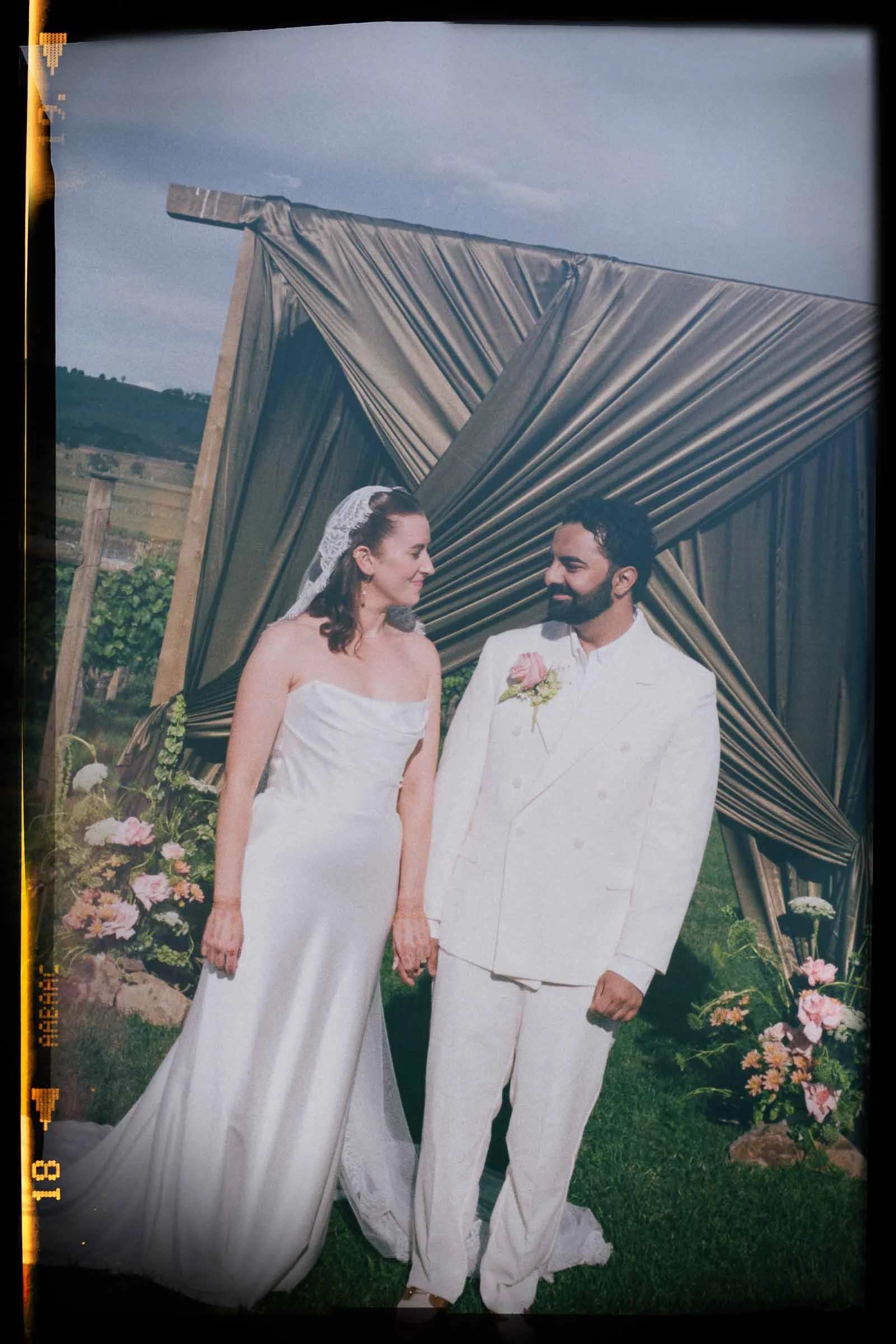A bride and groom holding hands during their wedding ceremony outdoors, standing beneath a decorative arch with draped fabric and flowers, with a cloudy sky above.