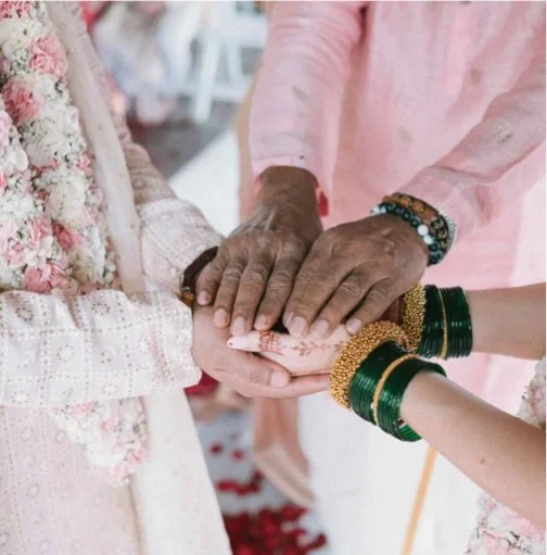 Person in traditional attire holding hands during a ceremony, with others touching their hands, wearing colorful bangles.