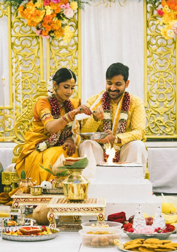 Indian bride and groom participating in a traditional wedding ceremony, seated at an altar decorated with flowers and gold accents, sharing a ritual.