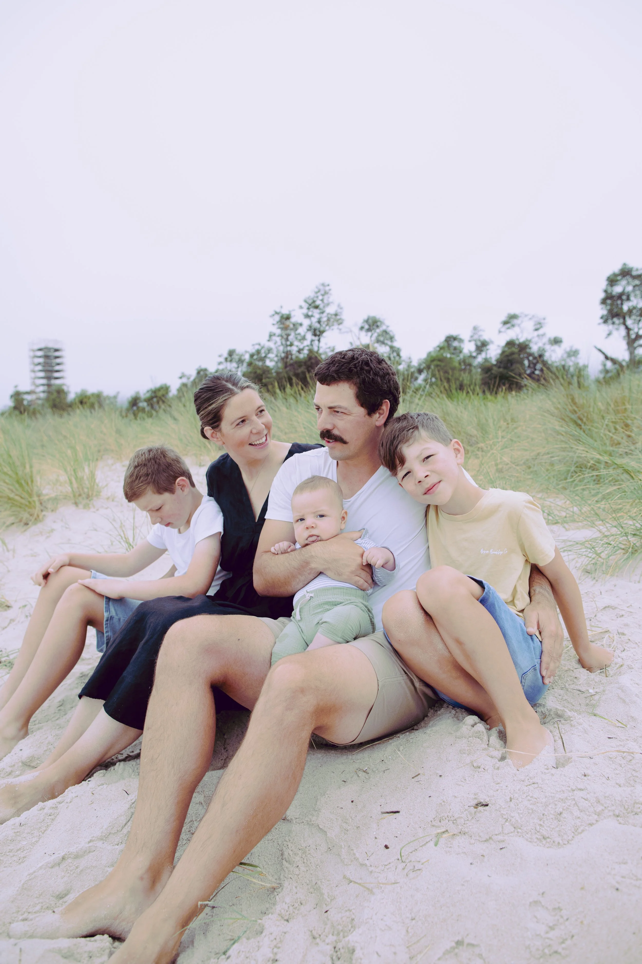 A family sitting on the sand at the beach with grassy dunes and trees in the background, enjoying a day outdoors.
