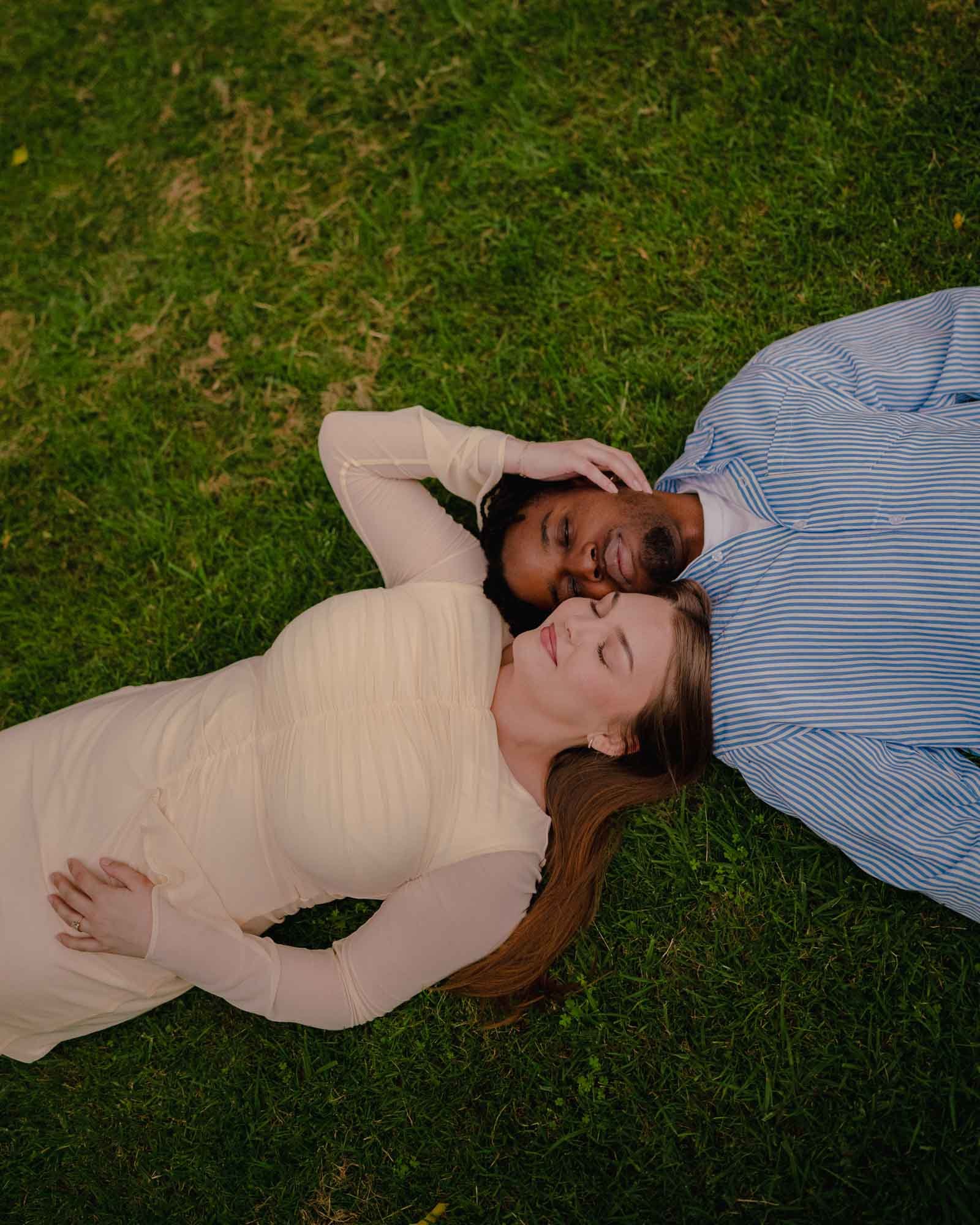 A man and a woman lying on grass, facing each other, with the woman resting her hand on her stomach and the man touching his head, both smiling with their eyes closed.