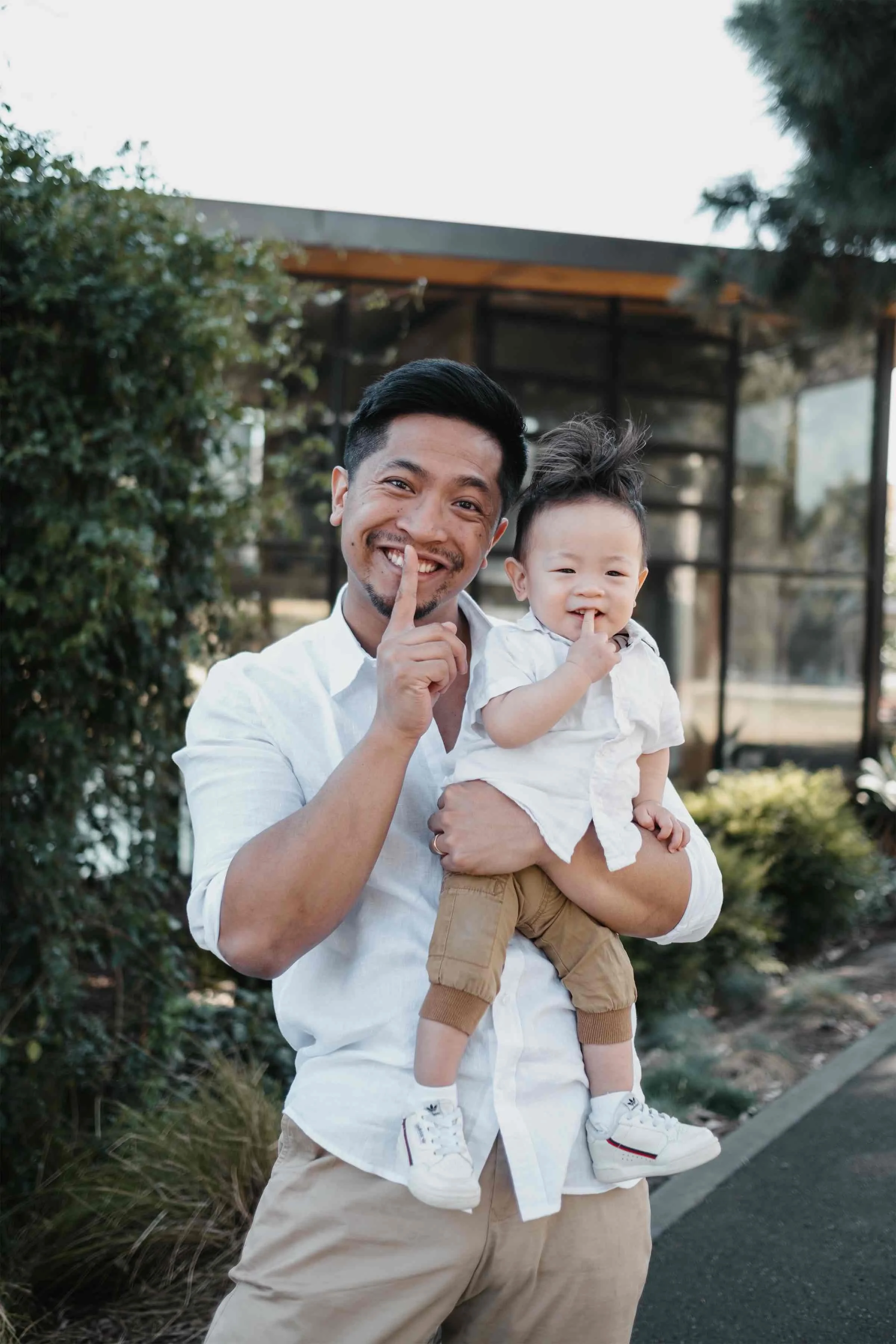 A man with dark hair and a beard holding a smiling toddler with dark, spiky hair. Both are making a finger to their lips gesture. They are outdoors, with greenery and a modern building in the background.